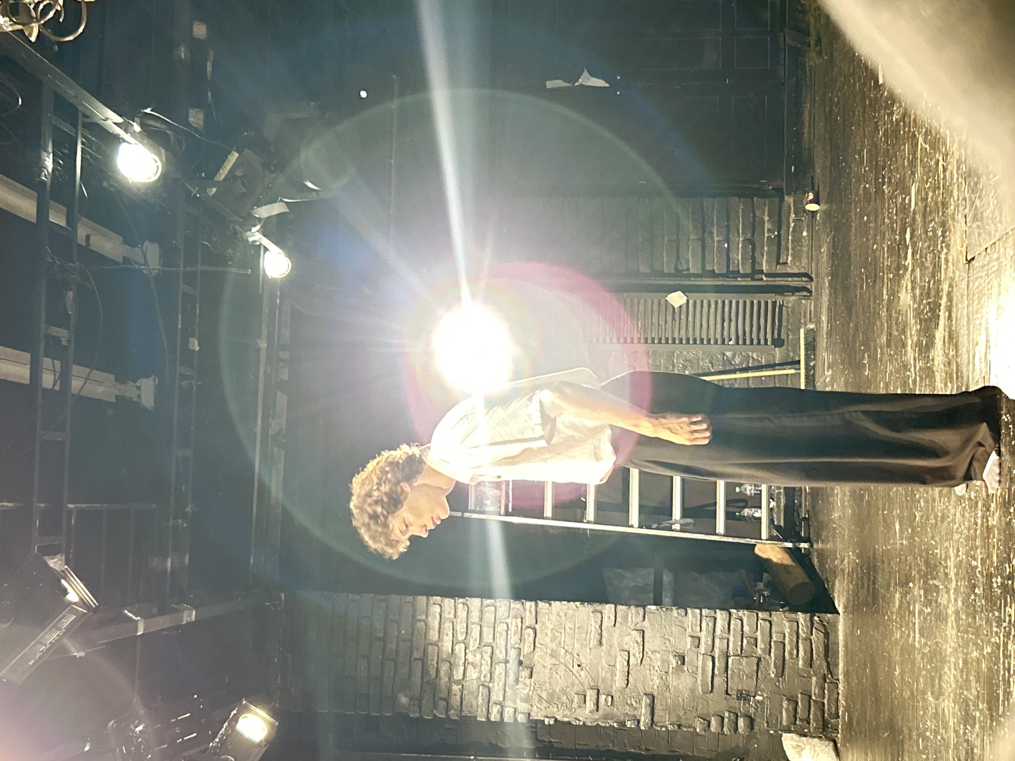 A young, slender male with curly brown hair stands on a wooden stage, looking down and to his right. He is wearing a white, short-sleeved collared shirt and loose-fitting black pants, with grey socks visible. The background features a brick wall painted black, a tall silver ladder, and a set of closed wooden doors. Stage lights are visible overhead, creating lens flares in the image. The stage floor is scuffed and appears to have some debris. The setting is likely a theater or performance space in an unknown location.