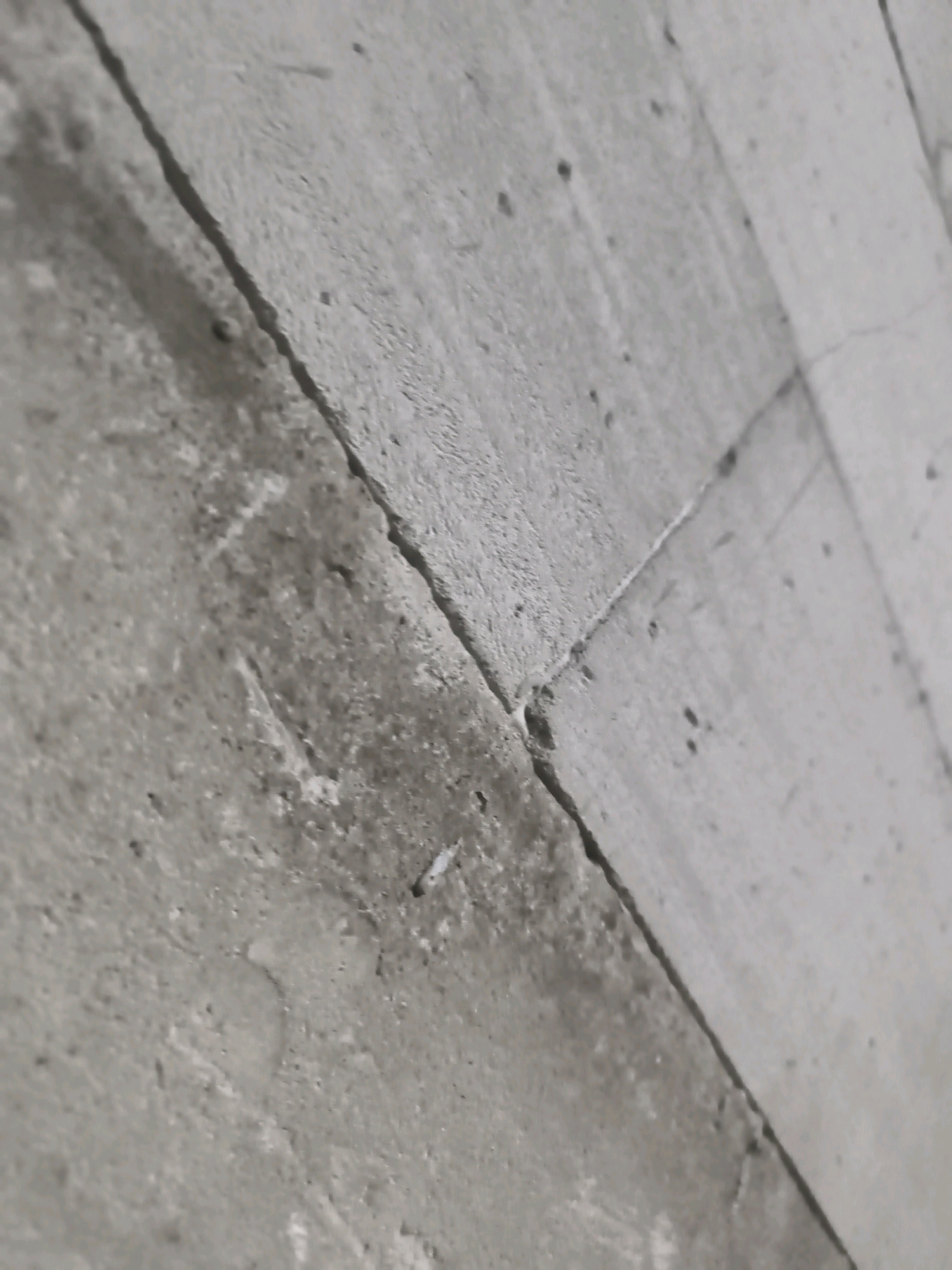 The image shows a close-up view of a concrete surface with a visible crack running diagonally across it. The concrete appears aged and textured, with some small dark spots and variations in shade. The lighting suggests an indoor or shaded outdoor environment, casting subtle shadows. There are no people or any other distinguishing features visible in the frame. The location of Sumogawe, Indonesia is not discernable from the visual information provided.