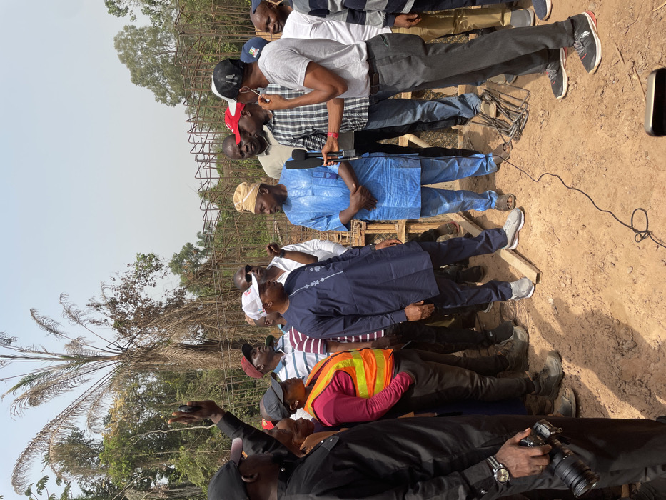 A group of men stand outdoors, observing something out of frame. Some wear traditional African clothing, including a light blue top and brown cap. One man wears an orange safety vest. Another holds a camera. Several trees are visible in the background, along with a structure made of steel rebar. The ground is reddish-brown, suggesting a dirt or clay surface.