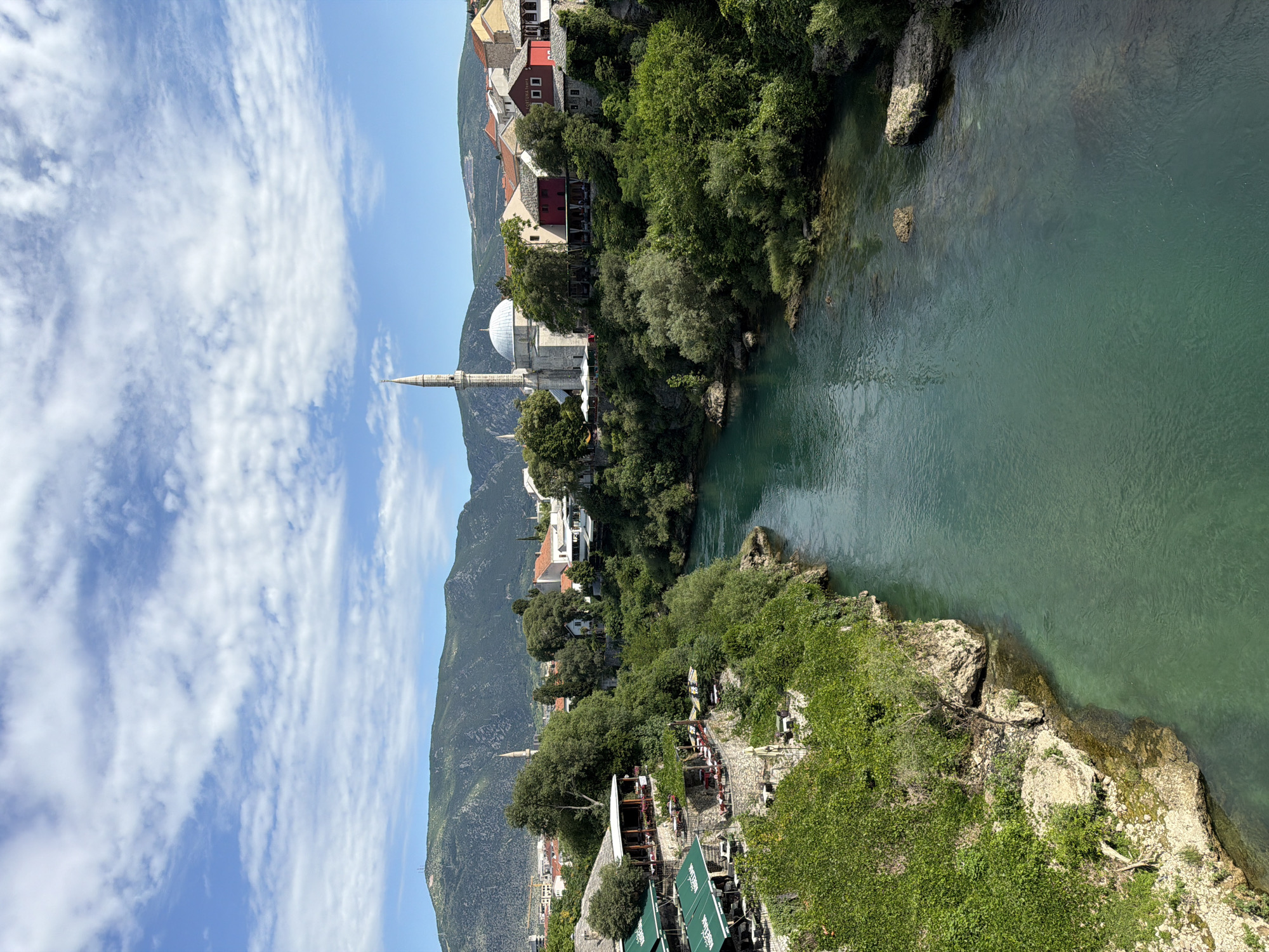 The image presents a panoramic view of the Neretva River in Mostar, Bosnia and Herzegovina, flowing through a verdant gorge under a partly cloudy blue sky.
The upper portion of the frame features a bright blue sky with scattered white stratocumulus clouds. Below the sky, distant, rugged mountains covered in green vegetation span the horizon; one prominent peak on the left exhibits antenna towers.
The central and dominant feature is the turquoise-green river, which flows from the upper center towards the bottom of the frame. The water is clear, revealing the rocky riverbed in shallower sections. Both riverbanks are steep and extensively covered with dense green foliage, including trees, shrubs, and ivy clinging to the rocky terrain.
On the left bank, a series of terraced structures, identified as cafes or restaurants, descend towards the river. These establishments feature multiple levels of outdoor seating areas with tables and chairs, shaded by large green awnings, some with text visible (e.g., "KAJRNA" or similar). These terraces are supported by stone retaining walls integrated into the landscape.
On the right bank, a cluster of buildings characteristic of Mostar architecture is visible. A prominent white mosque with a tall, slender minaret and a large dome stands out, surrounded by several multi-story buildings with red-tiled roofs and facades in red, white, and stone. One building displays the text "FORTUNA-TOURS." These structures are interspersed with trees and shrubs, and some also feature outdoor seating areas.
The scene is illuminated by bright daylight, casting clear shadows, indicating a sunny day. No discernible human figures are clearly visible. The perspective is from an elevated vantage point, looking downstream over the river and its banks.