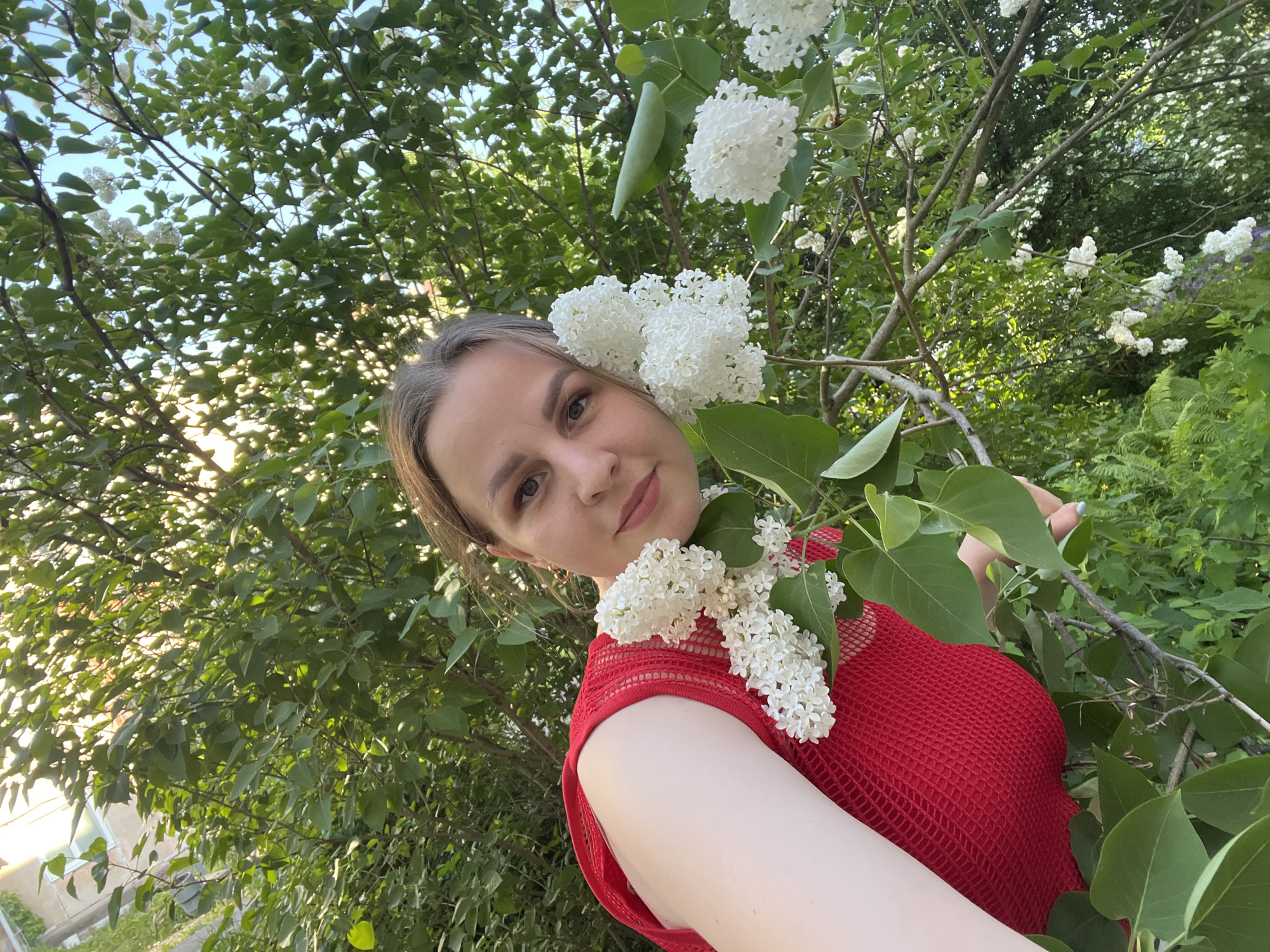 A young woman with light brown hair and fair skin is posing for a selfie in front of a tree with white flowers. She is wearing a red, sleeveless mesh top. The flowers are clustered near her head and shoulder. The background consists of green foliage and branches. The location appears to be outdoors, possibly a park or garden. There are no identifying features to confirm that this location is in Izhevsk, Russia.
