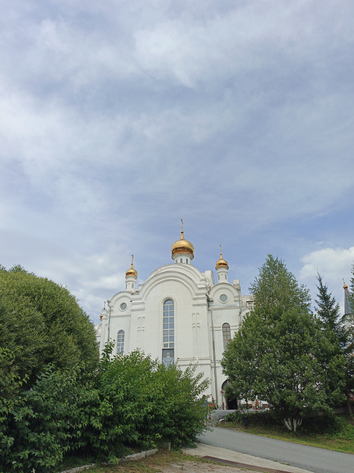This is a low-angle, full-frame, vertical shot of the White Orthodox Church in Zlatoust, Russia, under a partly cloudy sky. The church is a white, multi-domed structure with golden cupolas topped with crosses. It features arched windows and decorative architectural elements. Lush green trees and bushes partially obscure the lower portion of the church. A paved path leads towards the entrance, where several people are visible in the distance, blurred due to the distance and focus of the shot. The ground in the foreground is covered with dry grass and some concrete edging. The overall atmosphere is serene and majestic.