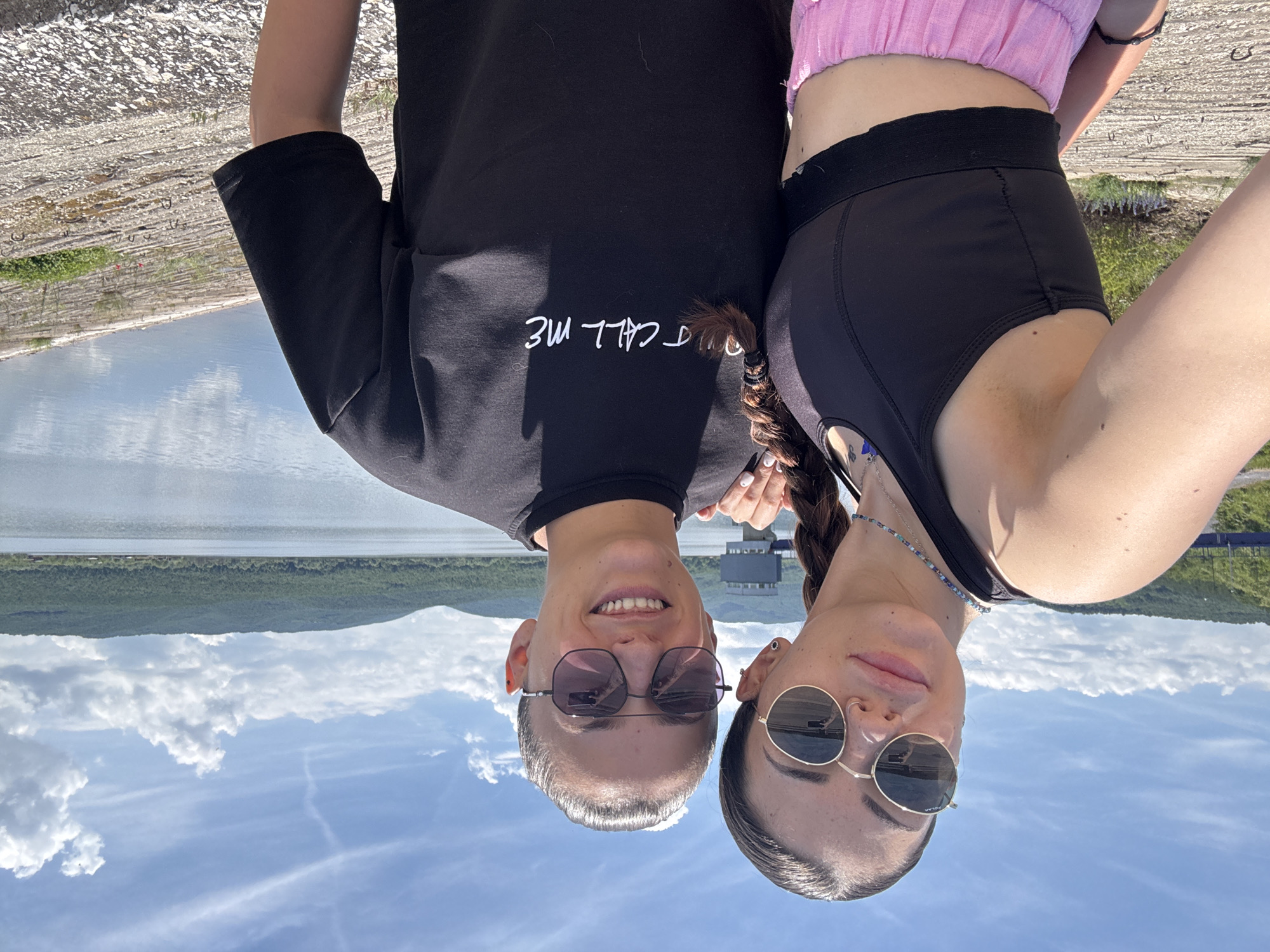 An outdoor portrait captures two young women posing playfully, likely for a selfie, set against a scenic backdrop. The image is oriented upside down, but when viewed correctly, the subjects are positioned in the foreground, with their heads tilted slightly upwards towards the camera.
The woman on the left (when correctly oriented) has short, light-colored hair and wears a black t-shirt with the text "DONT CALL ME" printed in white on the chest. She is also wearing dark, hexagonal-framed sunglasses and offers a subtle smile. The woman on the right has long, dark braided hair, round sunglasses, and a neutral, relaxed expression. She is dressed in a black sports bra or crop top beneath a light pink ruffled top and black shorts. A delicate beaded necklace is visible around her neck.
The background features a vast body of water, likely a reservoir or lake, stretching across the mid-ground. Beyond the water, a lush green, forested hillside or mountain range rises. The sky above is clear and blue with scattered white, fluffy clouds, suggesting a bright and sunny daytime setting. The immediate foreground, beneath the subjects, appears to be a concrete or rocky surface, possibly an embankment or dam structure. The overall scene suggests a pleasant day spent outdoors in a natural landscape, consistent with the Ognyanovo Dam and Reservoir area in Bulgaria.