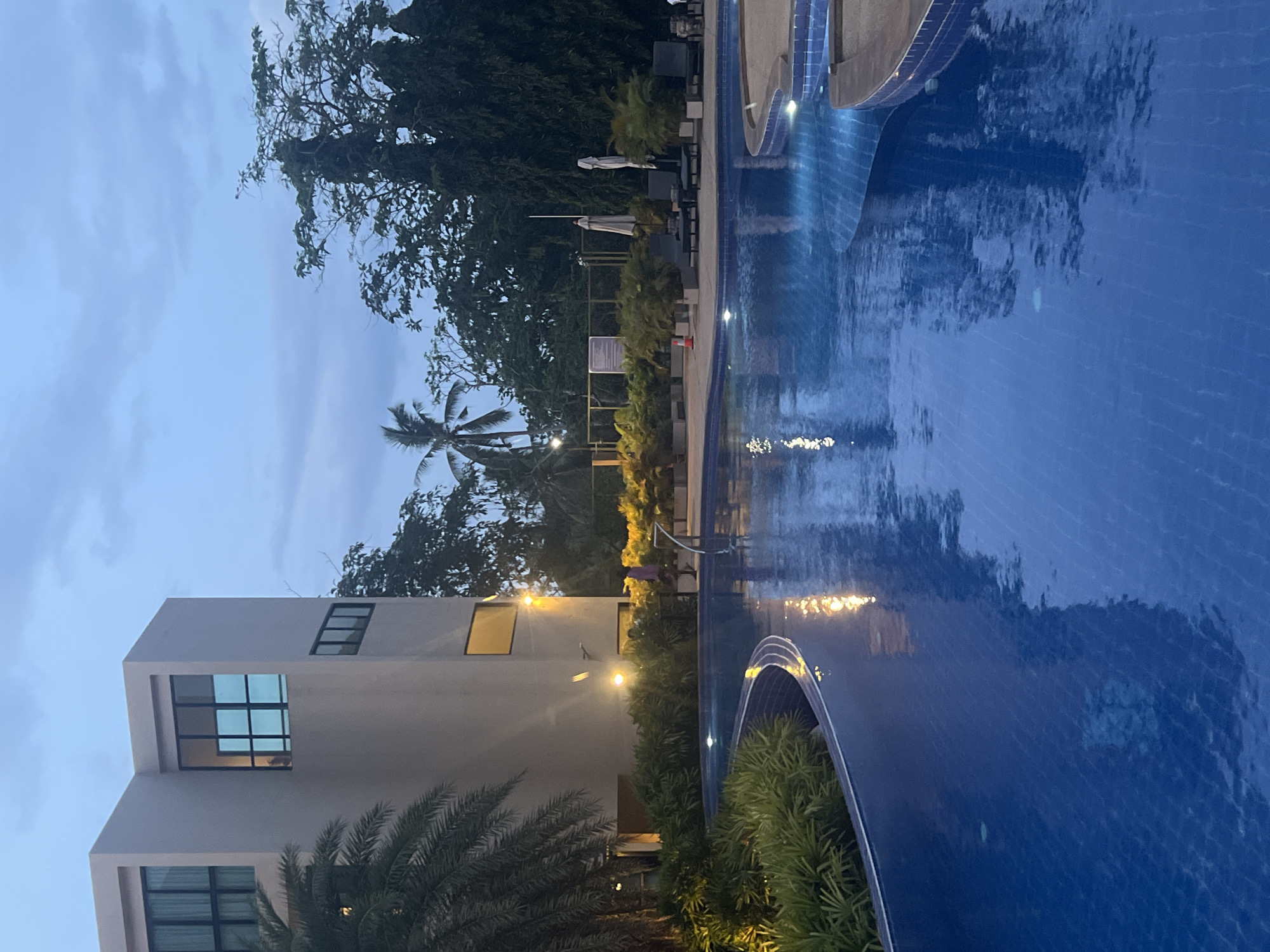 A poolside view at dusk in Ko Samui, Thailand. The image features a modern building with large windows, a palm tree in the foreground, and a curved swimming pool filled with blue water reflecting the evening sky and lights. Several lounge chairs with umbrellas are visible on the pool deck, surrounded by lush green plants. A person in a pink garment is walking near the pool. The sky is overcast with shades of blue and grey, indicating twilight. The overall scene depicts a tranquil resort atmosphere.