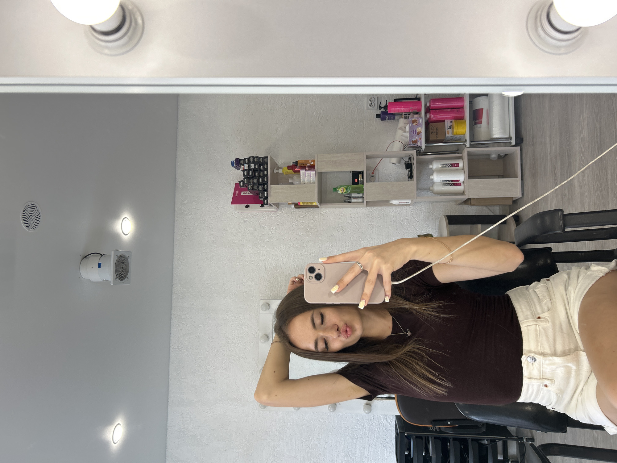 In a brightly lit room, a young woman with long brown hair sits in a black chair, taking a mirror selfie with her phone. She is wearing a dark brown t-shirt and white denim shorts. Her left arm is raised behind her head, and she is puckering her lips. The room appears to be a salon or dressing room, with a large mirror, shelves stocked with beauty products, and professional lighting. The walls are white and textured, and the floor is light gray wood laminate. A ceiling fan is visible in the background.