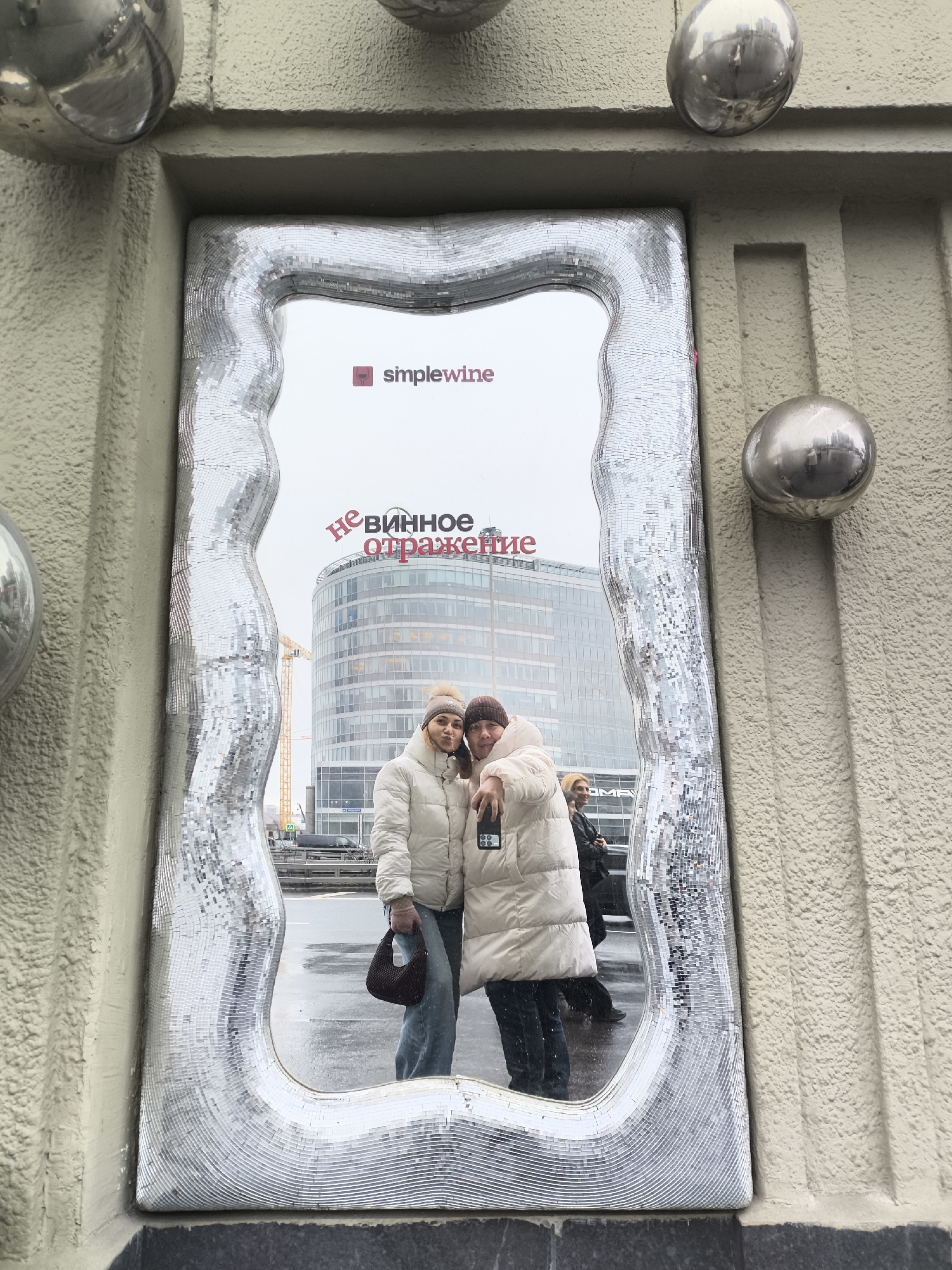 The image depicts two women reflected in a large, irregularly shaped mirror with a mosaic frame, set against a building's facade. The women, both wearing winter coats and hats, appear to be taking a selfie. One holds a phone, while the other poses slightly. Behind them in the mirror is an urban scene with a modern building. The building's signage indicates that it's located in Moscow, Russia. The exterior of the building with the mirror is textured and features decorative silver spheres. The environment suggests a cold, possibly overcast day. The sign on the building reads "simplewine" and "не винное отражение".