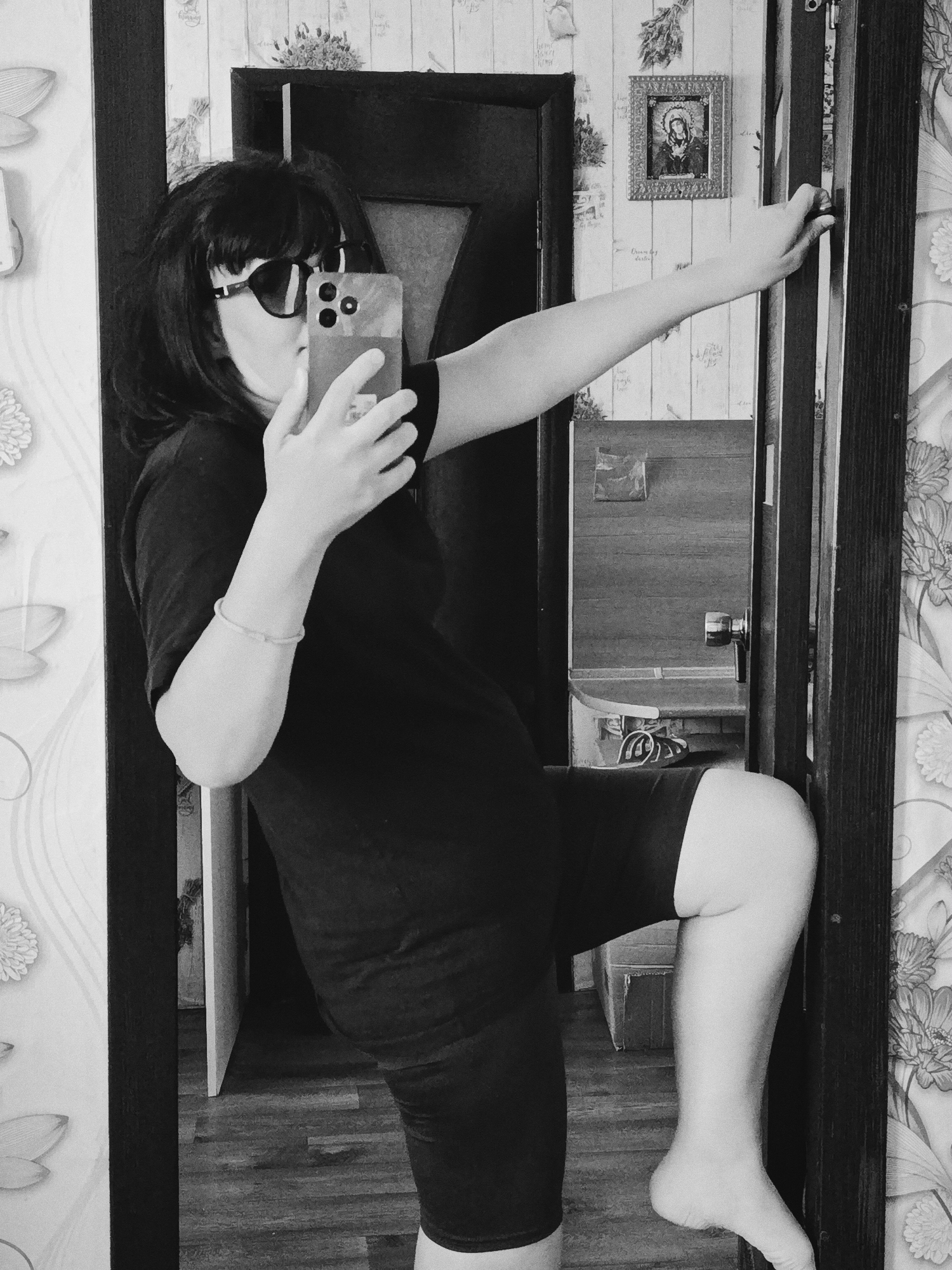 A person is photographed in a mirror, holding a smartphone to take a selfie. The person is wearing sunglasses, a t-shirt and shorts. Their left leg is raised, and their left foot is bare. The person is holding onto the door frame with their right hand. The background features wallpaper with a floral pattern and a framed picture on the wall. The location is Unknown, Unknown.