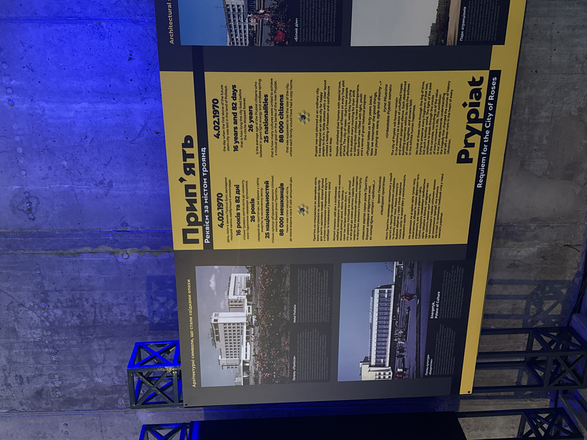 An informational exhibit about the city of Prypiat is prominently displayed in a dimly lit indoor space, likely a museum or exhibition. The poster, oriented vertically, is rectangular with a yellow bottom section and a black top section containing text and images. The background features a rough, textured concrete wall illuminated by a blue light, creating a stark contrast with the well-lit poster. Dark, geometric metal structures are visible in the foreground.
The exhibit's main title is "Прип'ять" (Prypiat) with the subtitle "Реквієм за містом троянд" (Requiem for the City of Roses), presented in both Ukrainian and English. Key dates and statistics are highlighted: "4.02.1970" (the day the first stone of the atom-city was laid), "16 years and 82 days" (duration of the city's life), "26 years" (average age of citizens), "25 nationalities," and "88 000 citizens" (planned population).
The text describes Prypiat as an ideal city for happy people, known for its wide streets, feeling of freedom, and citizens who cherished their homes, planting thousands of flowers, leading to its nickname "City of Roses." It touches upon the city's aspirations, youth, and sense of historical progress before describing the tragedy, focusing on unfulfilled hopes, poignant silence, and the echoes of past life in abandoned buildings. A quote from Iryna Pliusnina, "Unbeatable Prypiat women," is included.
The poster also features historical photographs under the section "Архітектурні символи, що стали свідками епохи" (Architectural symbols that became witnesses of the era), depicting buildings such as "Готель «Полісся»" (Hotel Polissia) and "Палац культури «Енергетик»" (Energetyk, Palace of Culture). Other sections, partially visible, mention "«Білий дім»" (White House) and "Парк атракціонів" (Amusement Park). The overall tone is melancholic, reflecting on the lost city and its history.