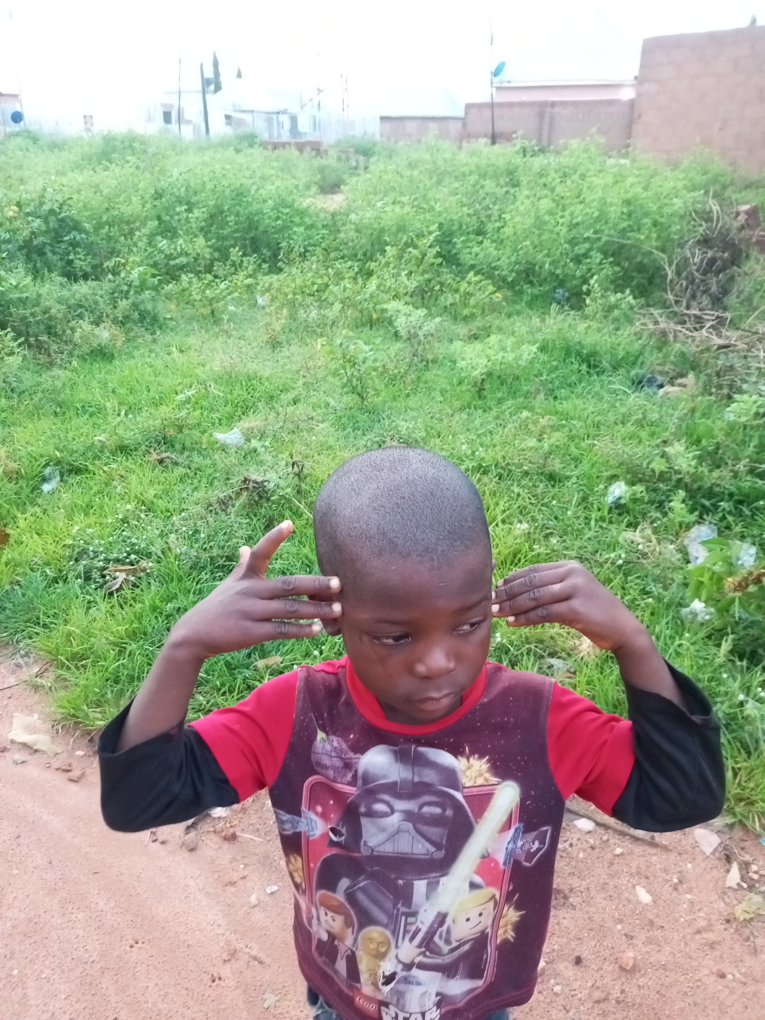 A young boy with a shaved head and dark skin stands in an outdoor, slightly overgrown environment. He is wearing a red shirt with black sleeves, featuring a print of Darth Vader and other characters from Star Wars. The boy is looking downwards and to his right, with his hands raised to his ears as if listening or blocking sound. The ground in front of him is dirt, and behind him is lush green vegetation. In the background, structures that appear to be buildings are visible, suggesting an urban or semi-urban setting. The sky is overcast and bright, indicating a cloudy day. The overall impression is a candid outdoor shot of a child in a somewhat unkempt environment.