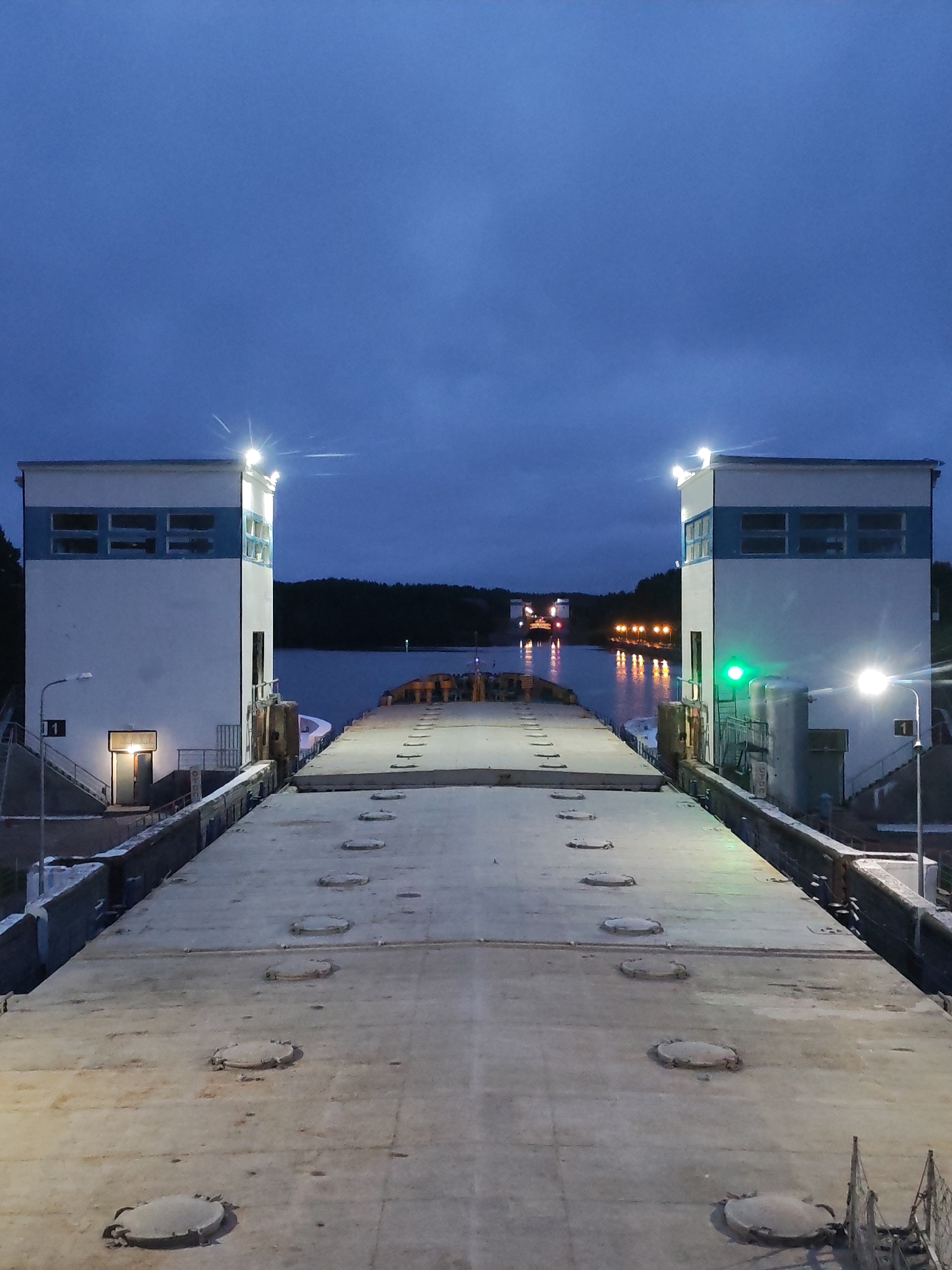 This image depicts the Markovo Lock, a waterway passage in Markovo, Russia, at dusk. The scene is dominated by a concrete lock chamber with two white, rectangular control buildings flanking its sides. These buildings have blue trim around their upper sections and multiple windows. Both structures are illuminated by bright spotlights on their roofs. The lock chamber itself is a wide, flat expanse of concrete, leading towards a body of water. Circular metal covers are embedded in the concrete at regular intervals. In the distance, a body of water is visible, with a ship or barge illuminated by lights, and further reflections of lights from other vessels or structures. The sky above is a deep blue, with some wispy clouds. A green traffic signal is visible on the right-hand control building.