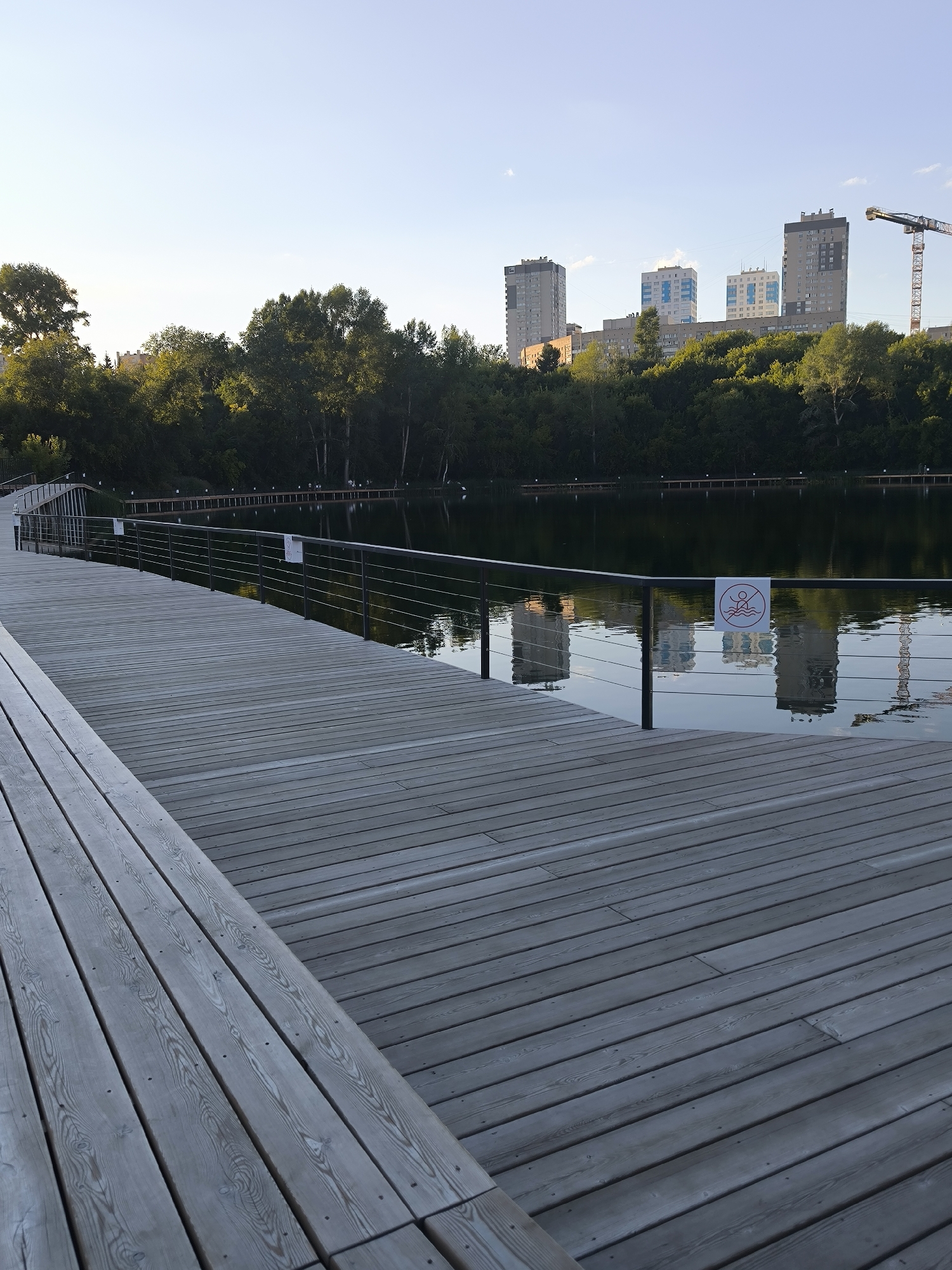 This is a photograph of a wooden pier and a body of water, likely a pond or lake, with a cityscape in the background. The pier is constructed of grey wooden planks laid horizontally and extends into the frame from the lower left. A metal railing with thin horizontal wires runs along the edge of the pier. On the railing, a circular sign with a red border depicts a stick figure in water with a red diagonal line through it, indicating "no swimming."
The water is calm and reflects the sky and the buildings behind it. A dense line of trees borders the far side of the water, with several tall apartment buildings and a construction crane visible behind the trees. The sky is clear and a pale blue, suggesting it is either early morning or late afternoon. The overall impression is one of a modern urban park setting. This scene is likely located in Novosibirsk, Russia, given the architectural style of the buildings and the presence of urban park infrastructure.