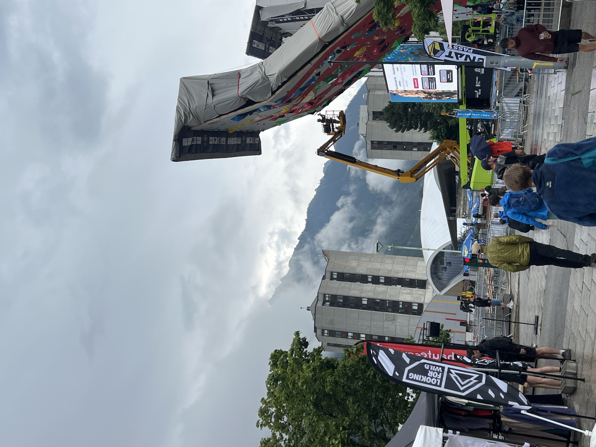 The image captures an outdoor event in Chamonix-Mont-Blanc, France, on a cloudy day. A large, multi-colored climbing wall dominates the right side of the frame, with a section covered by a gray tarp. A yellow boom lift is positioned near the climbing wall, with a person in the basket appearing to work on the wall. In the background, tall, concrete buildings and mountains shrouded in mist are visible. Various tents and stalls are set up in the foreground, with merchandise displayed. People are seen walking around the area, some holding umbrellas, indicating it may be raining. Flags and banners with logos are present, suggesting a sponsored event. A large digital screen displays advertisements.