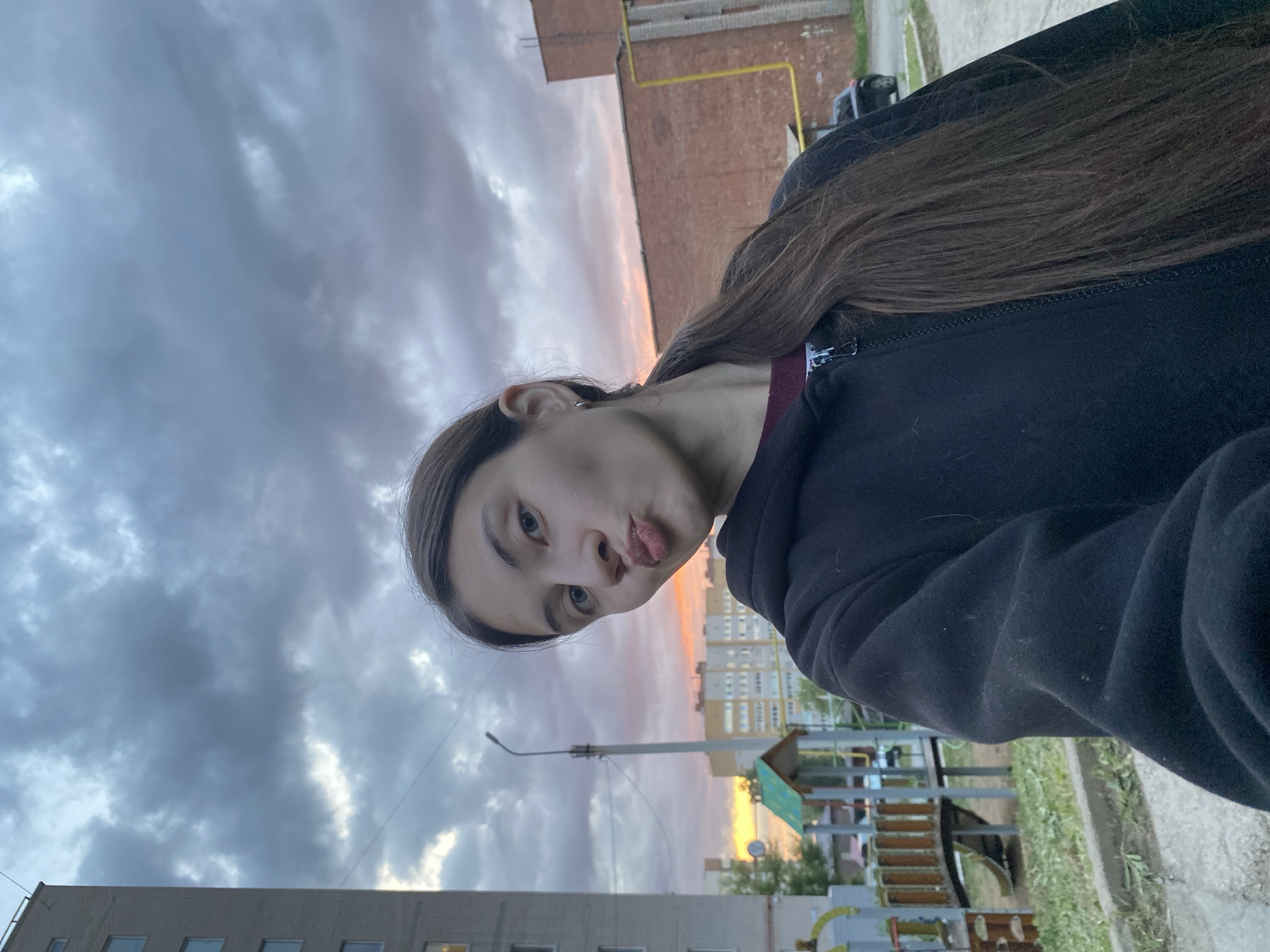 A young woman is taking a selfie outside, likely in the Russian city of Votkinsk. She has long, dark hair and is wearing a dark-colored hooded jacket. She is making a kissing face towards the camera. The background includes cloudy skies with a hint of sunset, apartment buildings, and a playground structure. A street lamp is also visible. The photo is taken at an angle, causing the horizon to appear tilted.