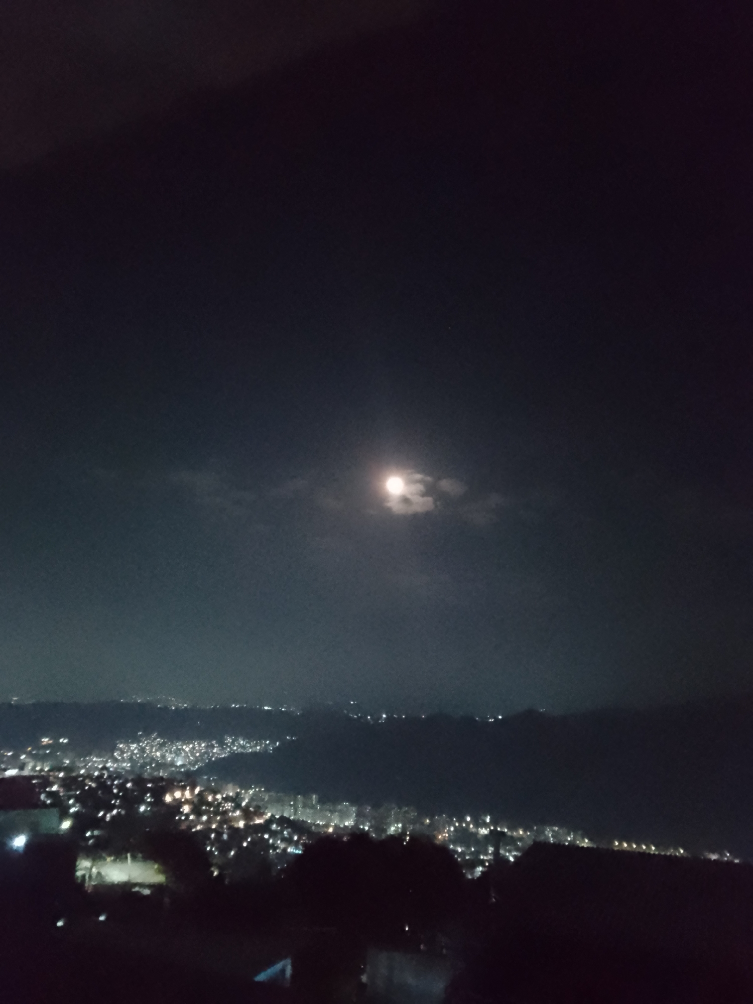 The image shows a night view of Caracas, Venezuela, with the city lights visible in the lower half of the frame. In the upper half, the night sky is visible with a full moon partially obscured by clouds. The foreground is dark, with some indistinct shapes of trees or buildings. No people are visible in the frame.