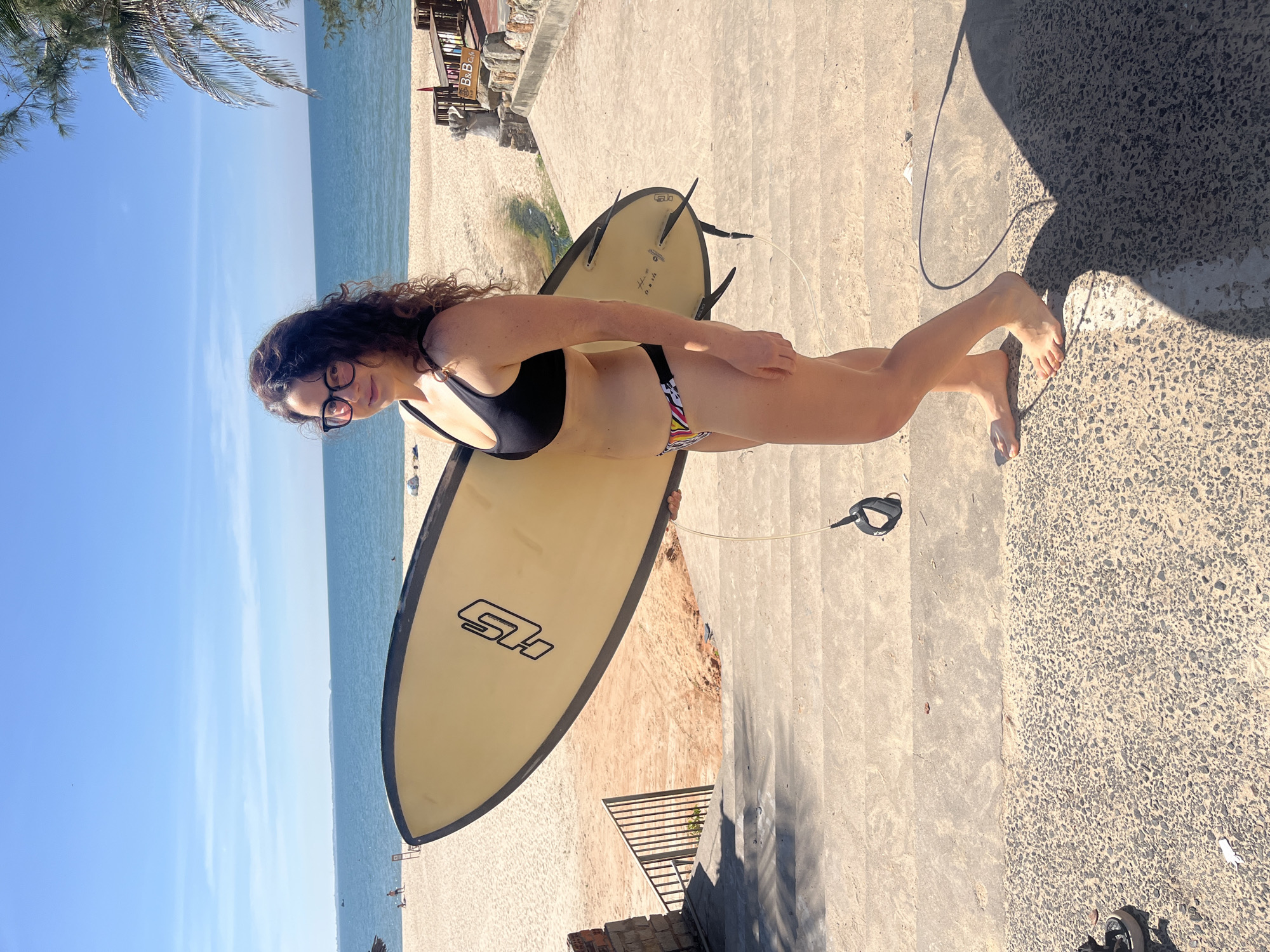 A woman in a black bikini stands on concrete stairs holding a yellow surfboard with black trim. She has curly dark hair and wears glasses. The surfboard has a black logo on it. Behind her, a sandy beach stretches to the ocean under a clear blue sky with some wispy clouds. There are some palm trees on the right side of the frame, and what appears to be a building with "B & B Cafe" signage on it. The scene is illuminated by bright sunlight, casting shadows.