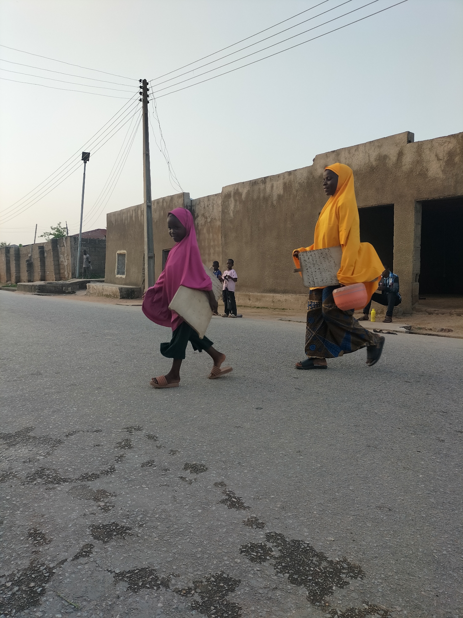The image captures a street scene in Jalingo, Nigeria, with two young girls walking across an asphalt road. The girl in the foreground, on the left, is wearing a pink hijab and dark pants. She is carrying a light-colored rectangular object under her arm and her feet are adorned with beige sandals. To her right, a slightly older girl, wearing a bright yellow hijab, dark patterned skirt, and black sandals, walks with a slight forward lean. She is carrying a wooden board with writing on it in one hand and a small orange object in the other. In the background, several other children are visible, as well as a man sitting down near an open doorway. The architecture suggests a developing urban environment, with unfinished concrete buildings lining the street. Overhead, power lines crisscross the sky, supported by a utility pole. The lighting suggests it is either early morning or late afternoon, with soft, diffused sunlight. The road surface shows some signs of wear, with patches of discoloration.