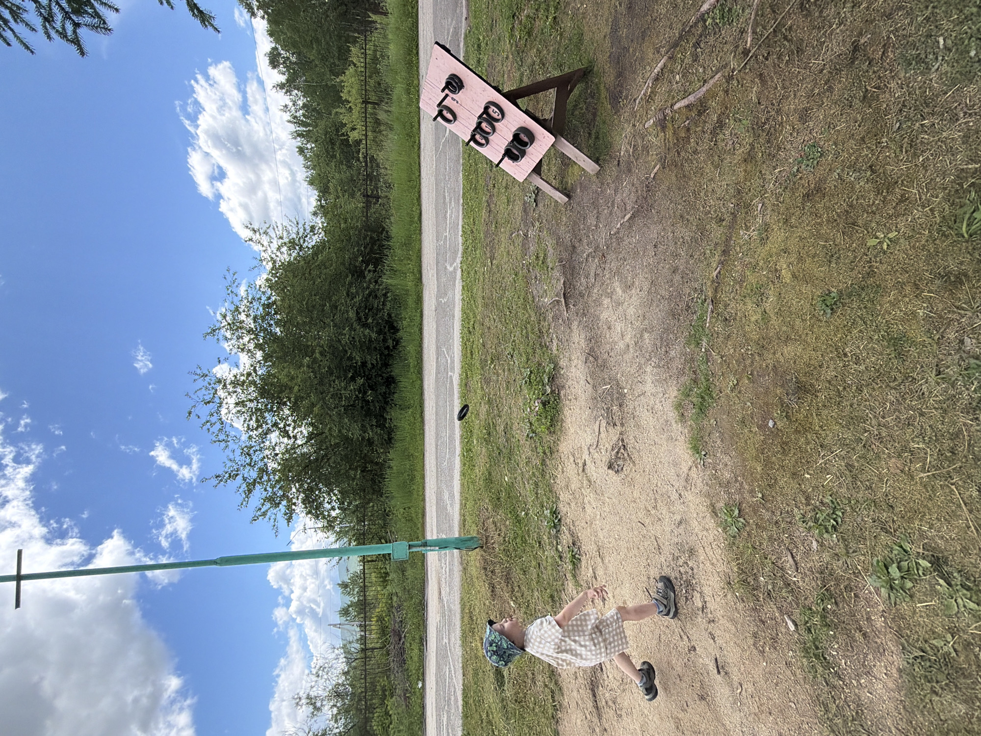 In the city of Kul'dur, Russia, a young child in a checkered outfit and a patterned hat is in mid-stride, about to throw a black ring. The ring is airborne, captured in motion. The child is on a dirt path, with dry grass and sparse green vegetation surrounding it. In the background, a road runs horizontally, bordered by more grass and dense green trees. Further beyond the trees, a chain-link fence and some indistinct structures are visible. A tall, green pole with a cross-shaped top stands on the left side of the image, near the child. To the right, a wooden target board stands on an easel, with several rings already placed on its pegs. The sky above is a vibrant blue, dotted with fluffy white clouds. The scene appears to be an outdoor recreational area.