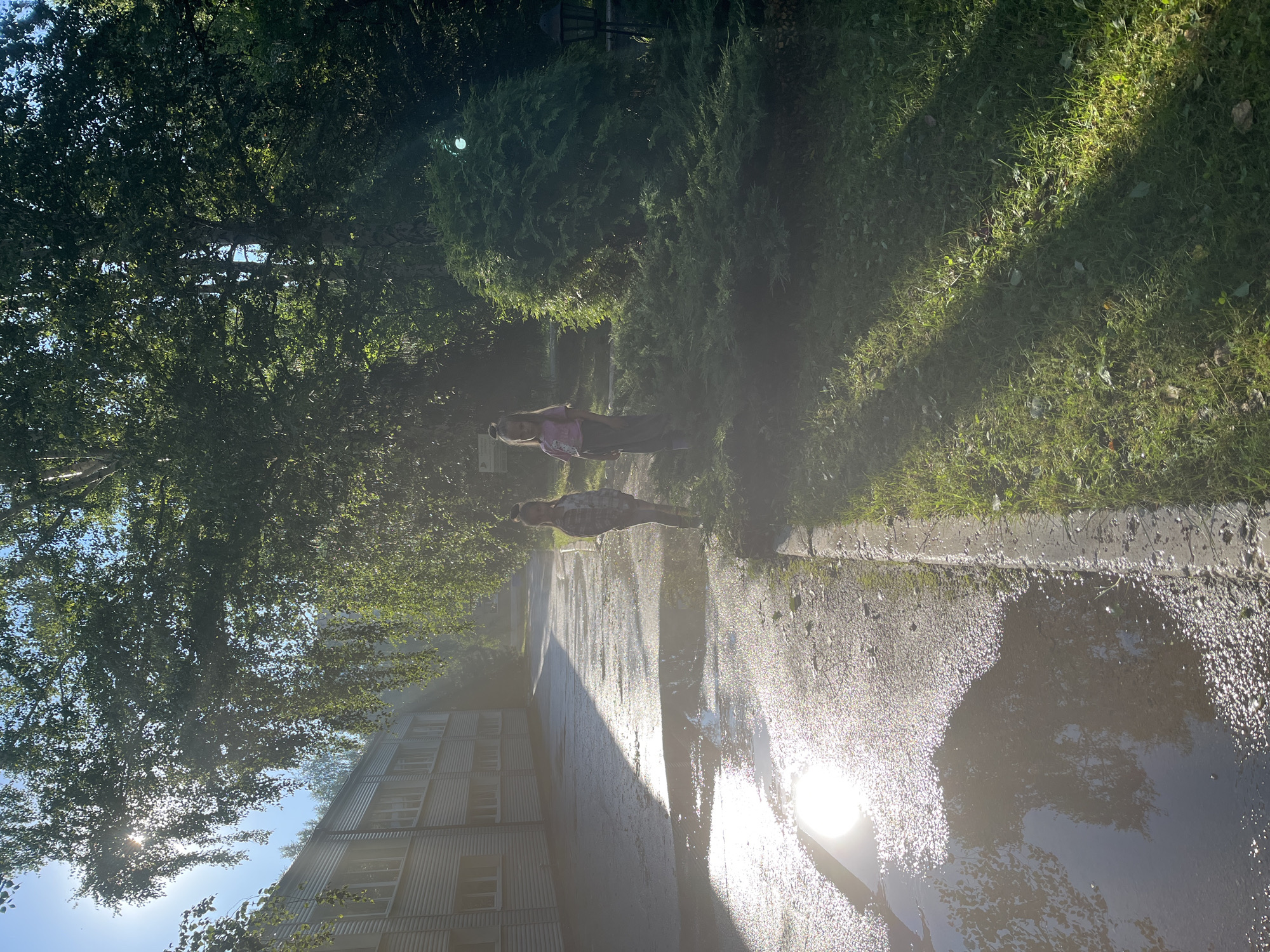 The image depicts a sunny outdoor scene, likely in Karagaiskii, Russia. The sun is bright, casting strong shadows and reflecting off a wet pavement or a large puddle in the foreground. The pavement is bordered by a grassy area on the right, and a line of trees and bushes on the left. In the background, a building with multiple windows and a corrugated metal facade is visible. Two individuals appear to be present in the scene, positioned between the trees and the grassy area. One person is wearing a pink shirt and dark pants, while the other is dressed in a checkered shirt and dark pants. Their presence suggests they might be walking or standing in this outdoor space. The overall impression is a bright, possibly humid day, with the sun's glare creating some lens flare and a hazy effect in certain areas of the image.