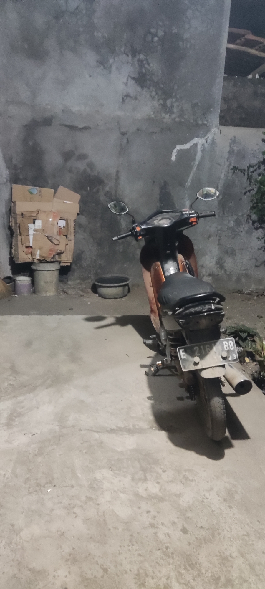 The image depicts a scene in the city of Karangwangun, Indonesia, under dim lighting conditions, likely during the evening or night. In the foreground, a weathered, orange-brown motorcycle is parked on a concrete surface. The motorcycle is facing away from the camera, with its rear visible. It has a black seat and a license plate that reads "E 2161 BB". To the left of the motorcycle, there are stacked cardboard boxes placed on top of a cylindrical container, and another smaller container to the left of that. Further to the left, a broom is leaning against the wall. Behind the motorcycle, a dark, circular basin is situated on the ground. The background consists of a rough, textured concrete wall, with some foliage visible on the right side of the frame. The overall impression is one of an outdoor storage or parking area.