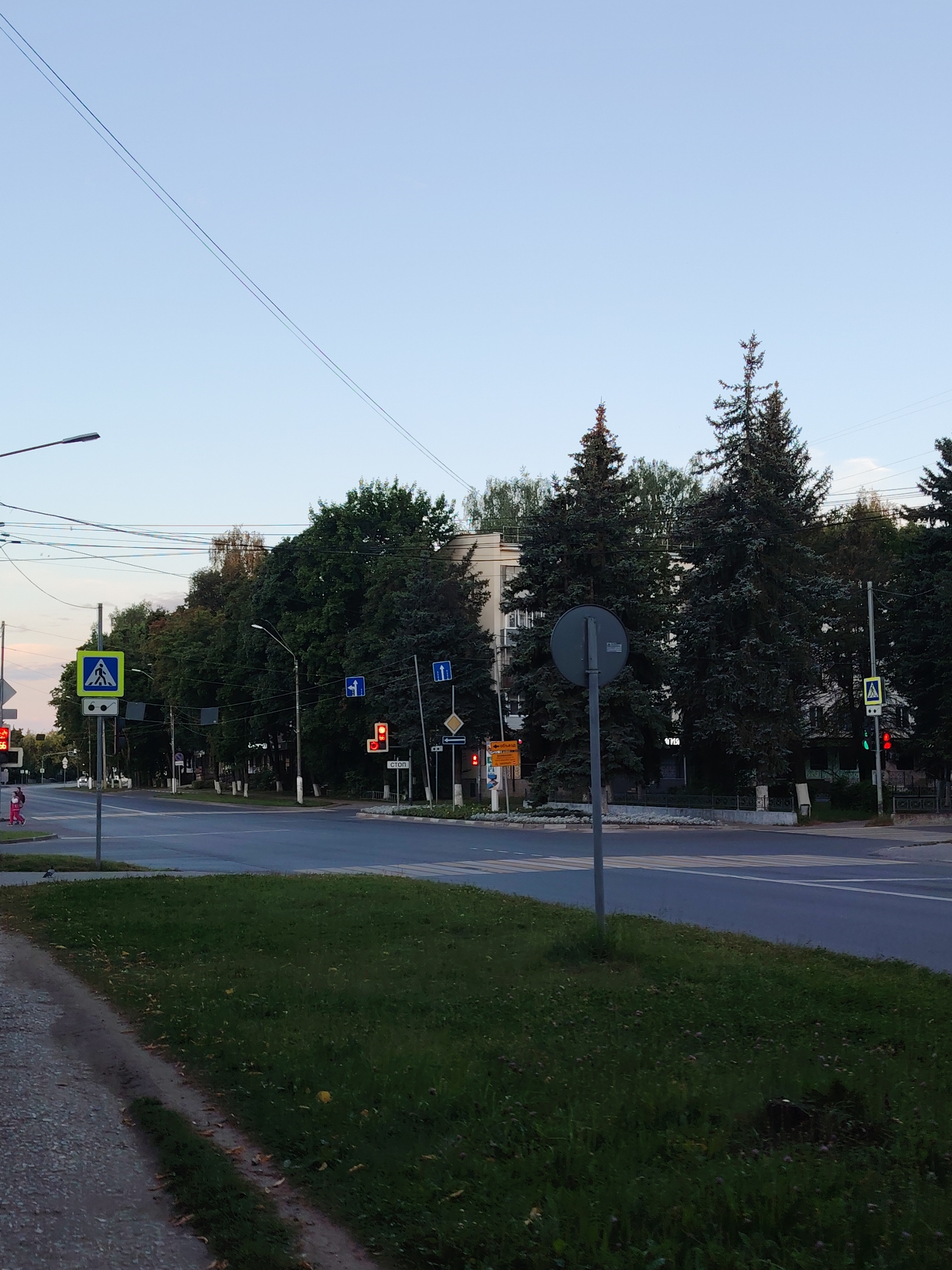 The image shows a street scene in Konakovo, Russia. It is daytime, with a clear sky. There is a road with traffic lights and pedestrian crossings. Several street signs are visible, including a pedestrian crossing sign and a stop sign. Tall evergreen trees line the street, and a multi-story building is visible in the background. A person in pink clothing is walking across the pedestrian crossing, and a traffic light is red, indicating that traffic should stop. A lamppost is also visible on the left side of the image. The grass verge on the right side of the road is well-maintained.