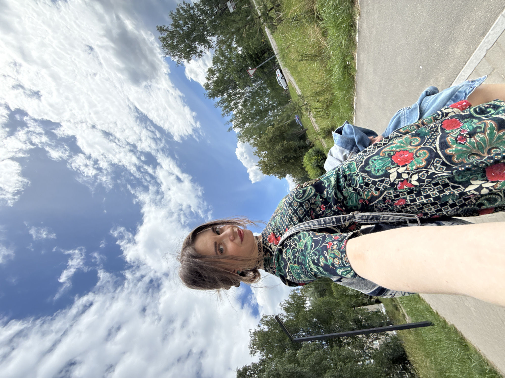 A young woman, with medium brown hair pulled back into a bun, is taking a selfie outdoors. She is wearing a dark, long-sleeved dress with a busy pattern of red flowers and green paisley designs, and a denim jacket. Her eyes are wide and looking directly at the camera, and she has a neutral expression. She is positioned in the foreground of the image, with a bright blue sky filled with fluffy white clouds behind her. To the right of the woman, there is a paved path, a grassy verge, and trees. A white car is parked on the side of the road in the background. A traffic sign with a red triangle and a white center is visible on a post among the trees. The scene suggests it is daytime, likely a sunny day due to the bright sky and clear visibility. The woman's clothing and the outdoor setting imply a casual, possibly recreational activity.