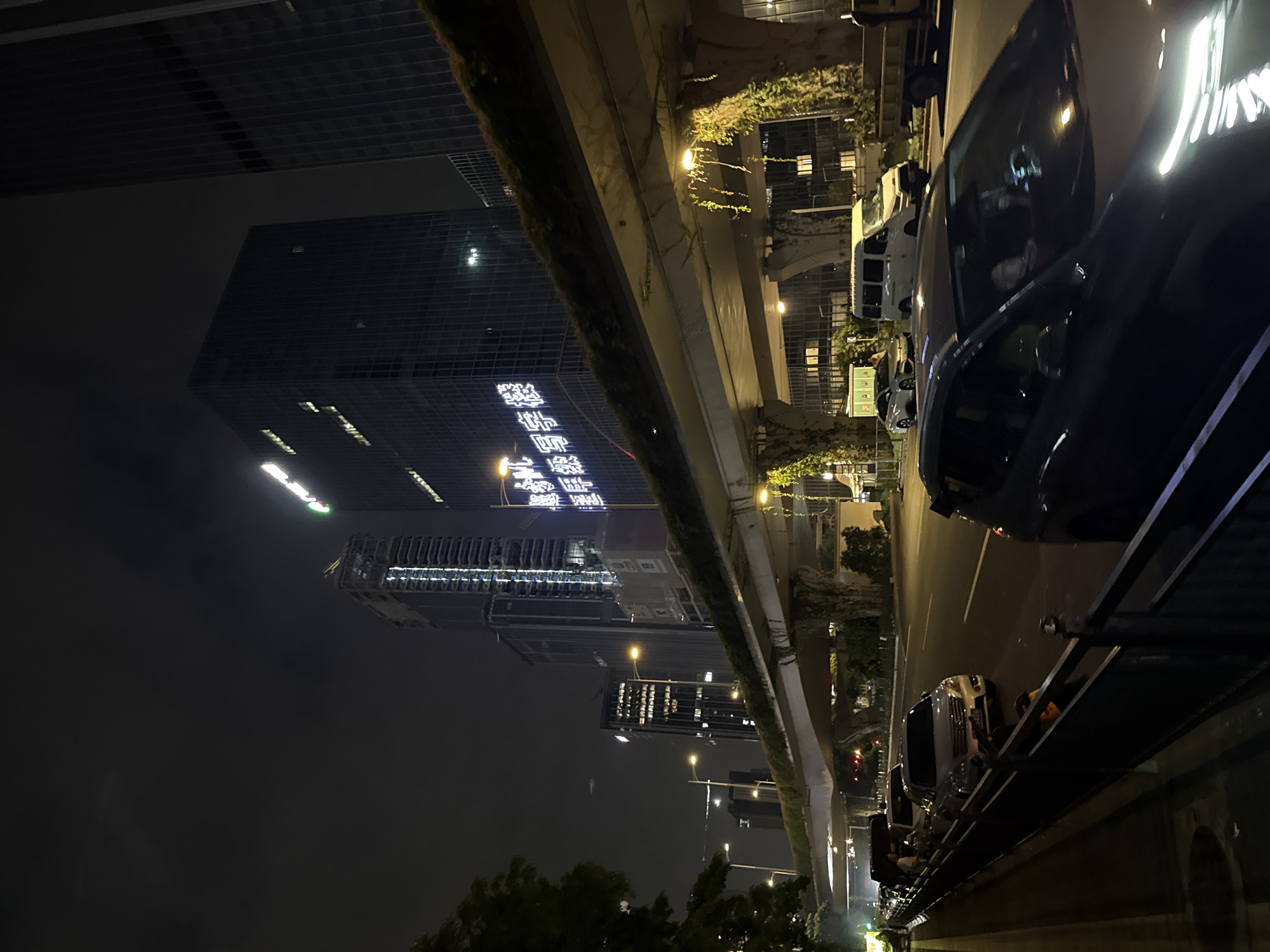 The image depicts a nighttime urban scene in Huangpu District, China, featuring a multi-level road infrastructure and modern architecture. A central focus is a section of elevated roadway covered in greenery, with vines trailing down its concrete supports. Below this elevated road, a street level features a line of cars, including a dark-colored sedan in the foreground with its headlights illuminated. Further down the street, several other vehicles are visible, some appearing to be in traffic. Tall buildings with illuminated windows dominate the background, with one prominent structure displaying bright white lettering in Chinese characters. The sky is dark, suggesting it is late at night, with visible streetlights casting a warm glow on the scene. A traffic light is visible in the distance.
