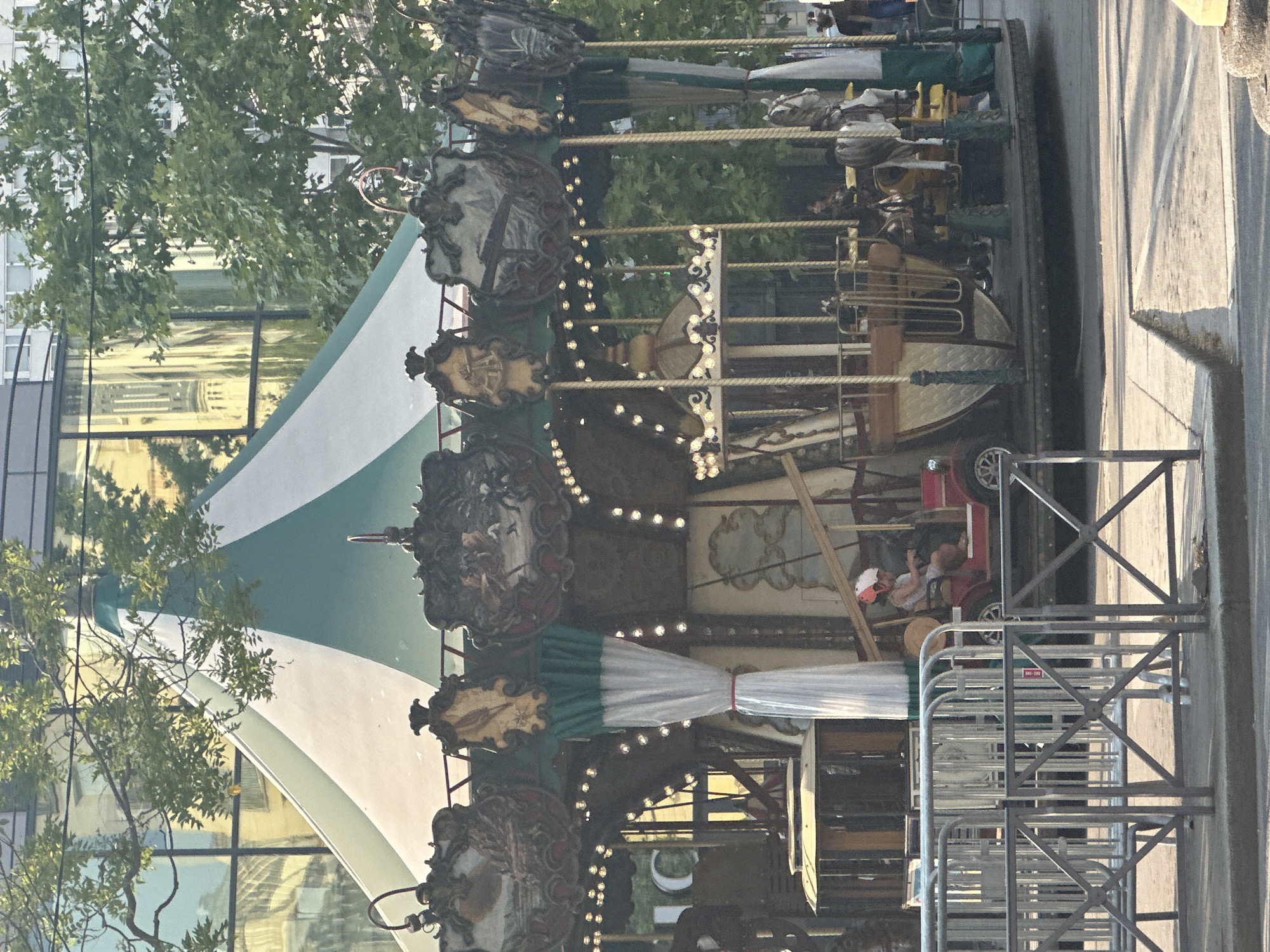The image depicts a carousel located in the Tuileries Garden in Paris, France. The carousel has a green and white striped canopy and is adorned with decorative elements and lights. Several carousel figures are visible, including horses and what appear to be small vehicles. In the foreground, a child wearing a helmet is seated in a red vehicle on the carousel. Behind the carousel, trees and a modern building with reflective windows are visible. The scene is outdoors, suggesting daytime.