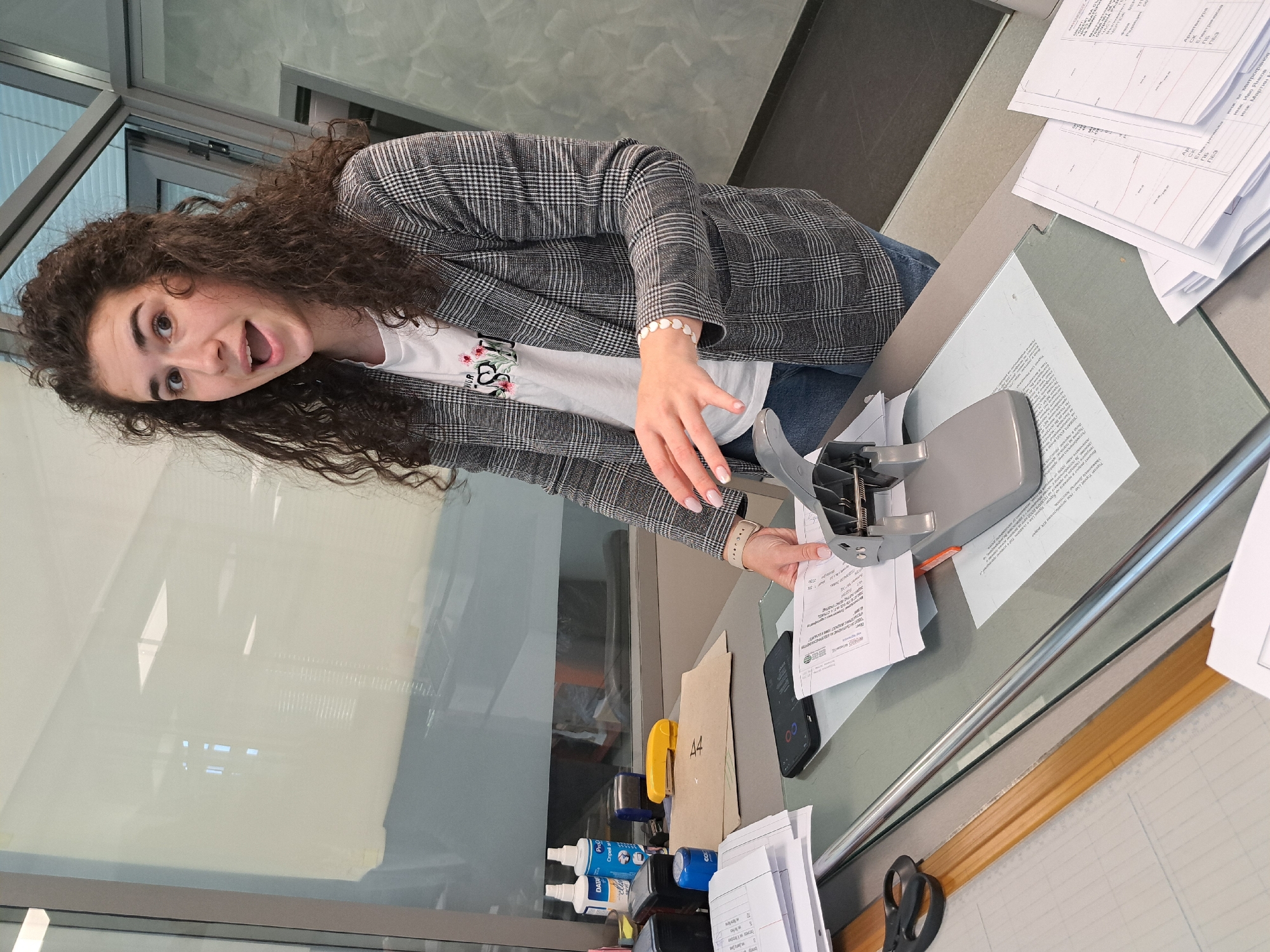 A young woman with long, curly brown hair is actively engaged with a large, grey paper punch on a desk, appearing to be in an office or administrative environment. She wears a grey plaid blazer over a white t-shirt with a floral print and blue jeans, with a pearl-like bracelet on her right wrist. Her mouth is open, suggesting she is mid-conversation or expressing surprise.
The desk has a grey surface and is partially bordered by a glass partition or window. On the desk, there are multiple stacks of white papers, some containing what appear to be architectural drawings or plans. A stack of papers is positioned within the paper punch. Other items on the desk include a black smartphone displaying "00," bottles labeled "Pro" and "Cnpell," a yellow stapler, a pair of black scissors, and a piece of cardboard marked "A4."
The background features a large window or glass wall, reflecting ambient light and indicating daytime. Text visible on the documents includes "DATA," "UR," "Ap," "BEP," and Cyrillic script such as "Архитектур," "CK," "Enew," "ПЕЗ," "инж. Мартин," "Иво Янков," and "иня м. митрофанов," suggesting a Bulgarian context, possibly related to engineering or architecture. The overall setting is a functional and bright workspace.