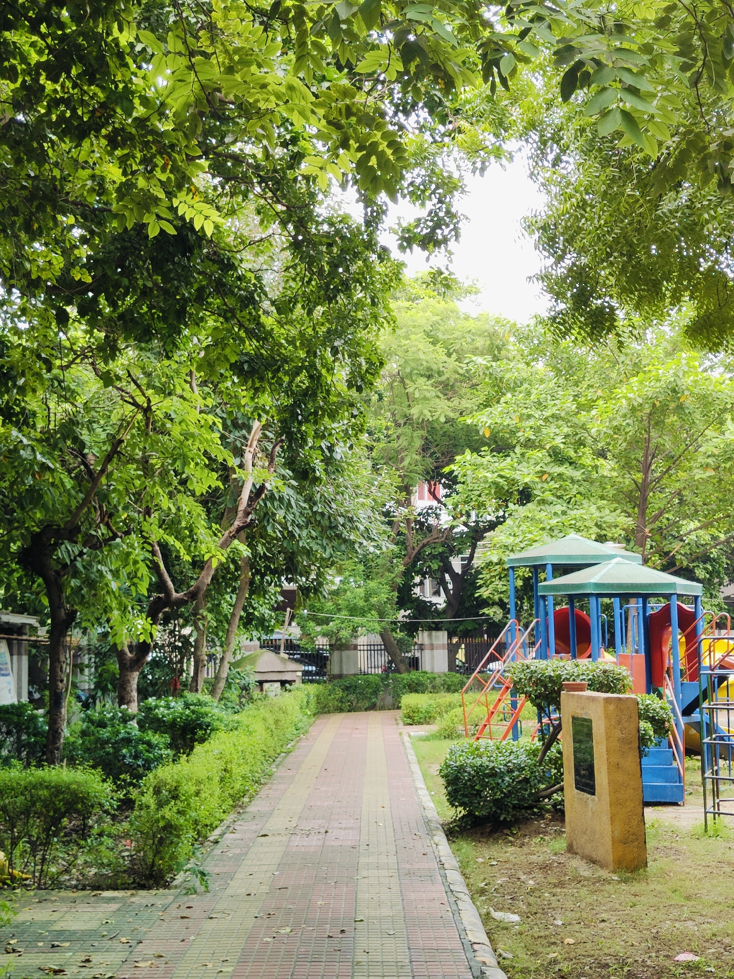 The image shows a paved walkway lined with manicured bushes and trees, leading towards a children's playground. The playground features a colorful structure with slides and climbing elements. A stone monument with an inscription is visible in front of the playground. The scene is set in a lush green environment, suggesting a park or residential area in Gurgaon, India. The sky is overcast, indicating a possibly humid or rainy day. There are no people visible in the image.