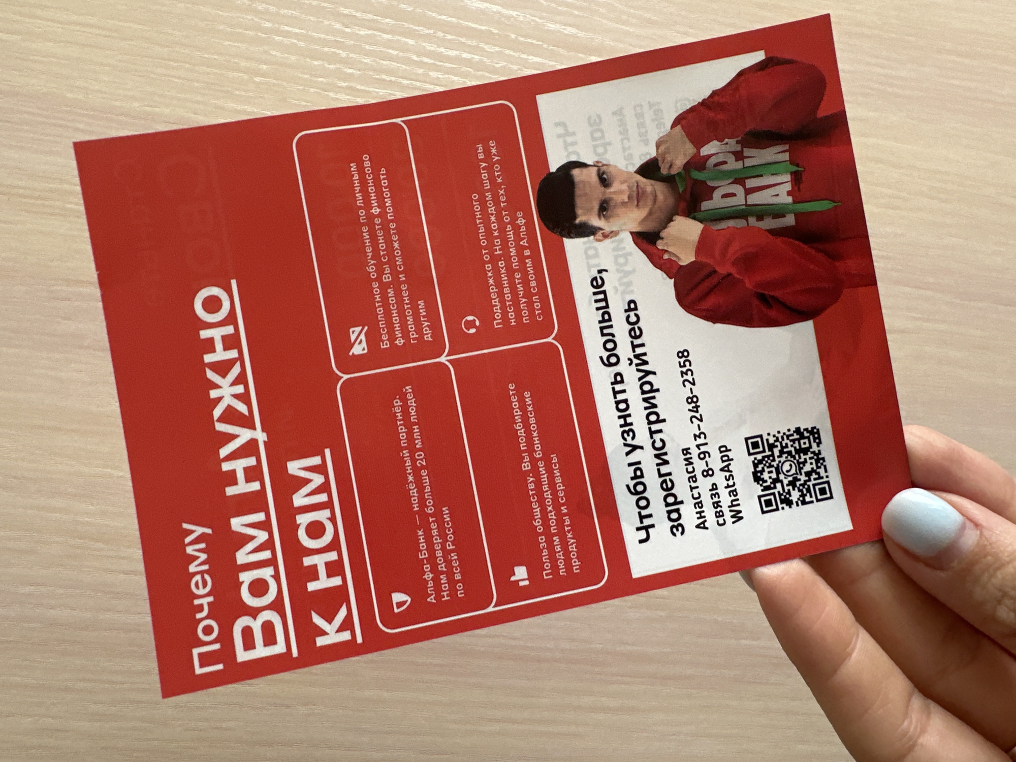 An overhead medium shot captures a red flyer held by a person's hand against a light-colored wooden surface. The flyer is mostly in focus, with the hand and wooden surface slightly blurred. The flyer features text and an image of a young man in a red hoodie. The text is in Russian and appears to be promotional. The hand, with a pale blue nail polish on one finger, is positioned to hold the flyer, obscuring part of the bottom right corner. The wooden surface suggests an indoor environment, possibly a table or desk. There is no identifiable information about the location being Novoaltaysk, Russia, or any specific city.