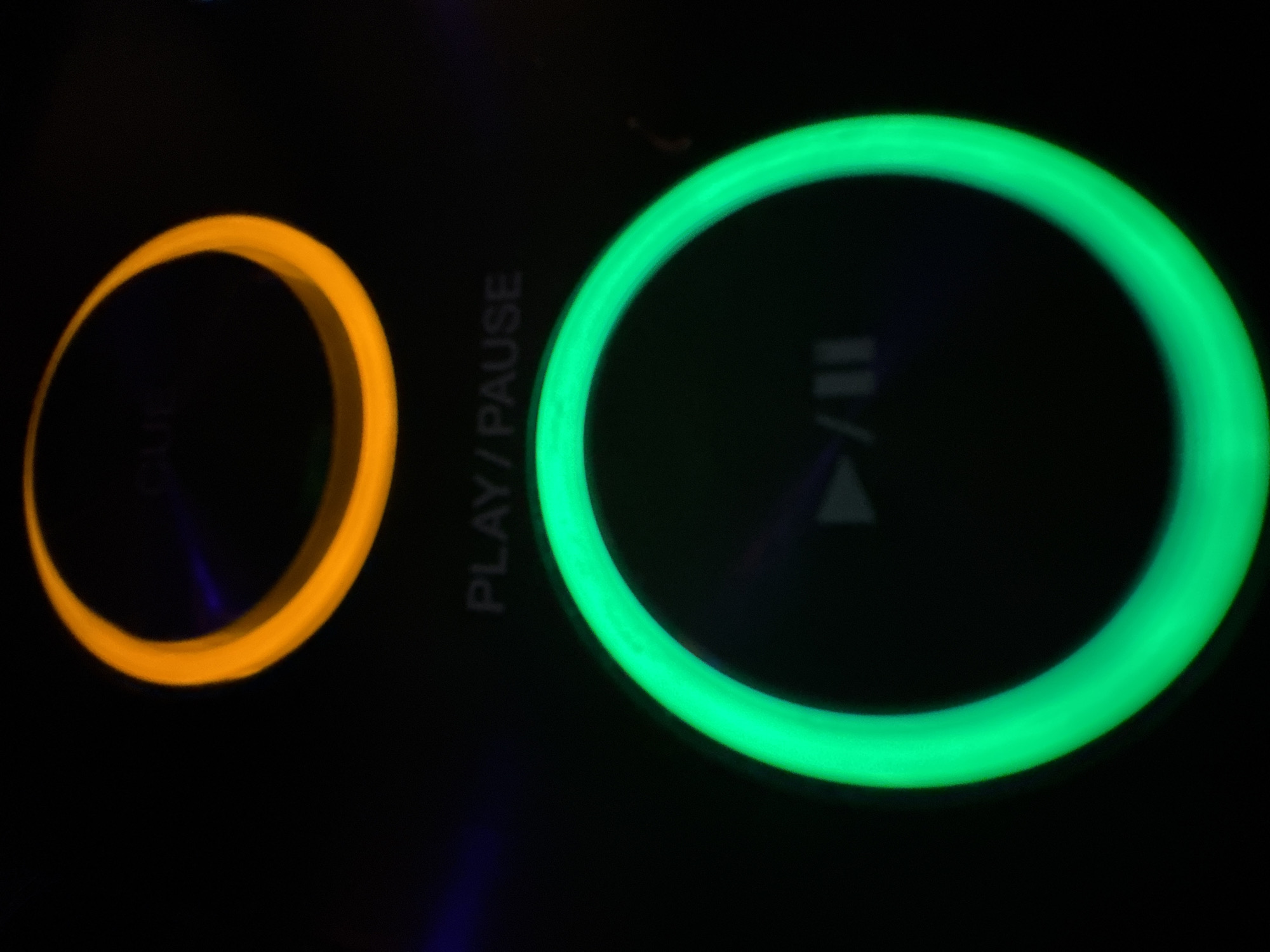 The image shows two illuminated circular buttons on a black surface. The left button is orange, and labeled "CUE", while the right button is green and labeled "PLAY/PAUSE" and contains a play/pause symbol. The buttons appear to be part of an electronic device. There is no information that suggests this image has any relation to Plovdiv, Bulgaria.