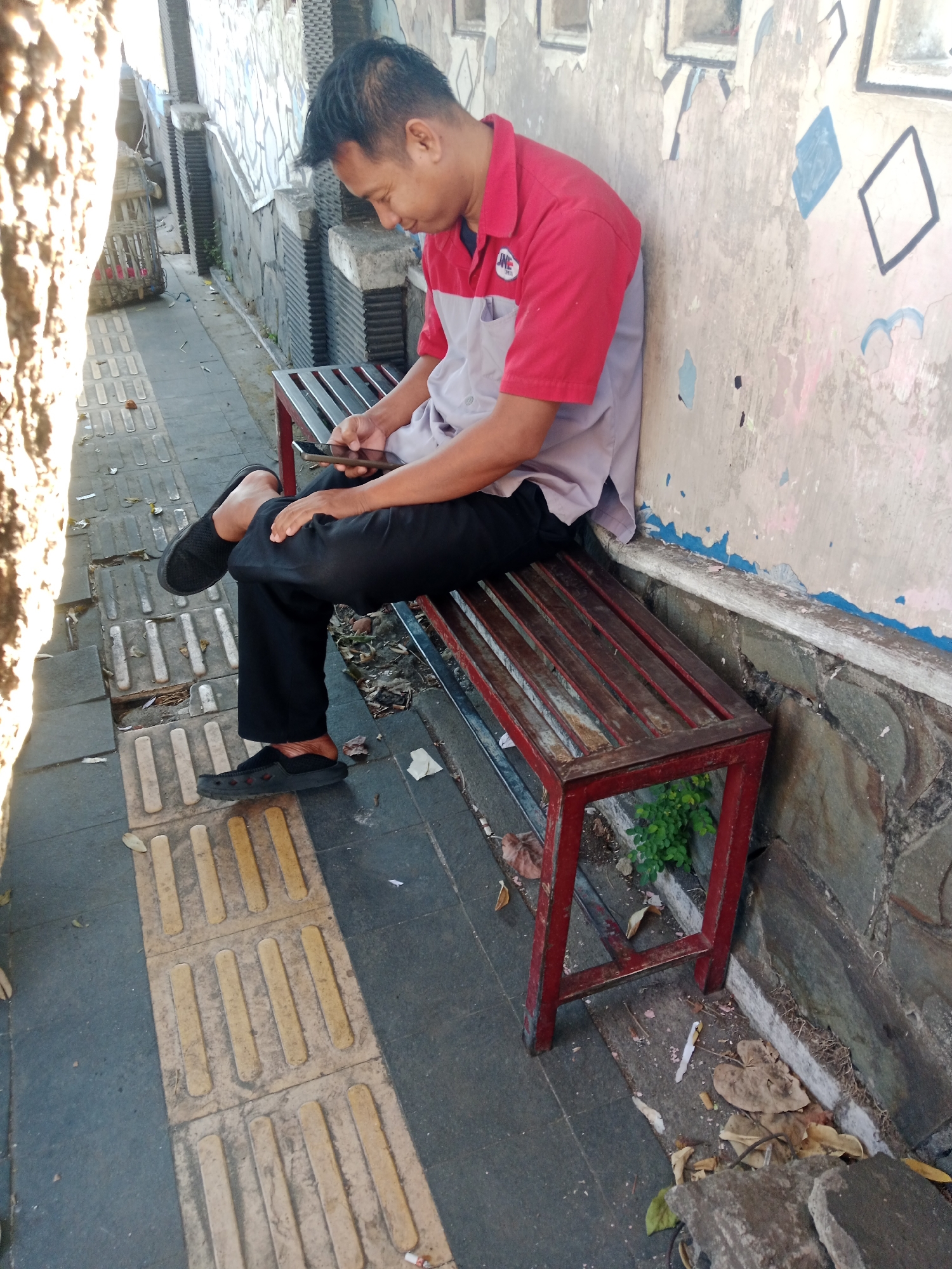 An East Asian man with short black hair is sitting on a red metal bench with wooden slats. He is wearing a red and grey polo shirt and black pants. The man is looking down at a black smartphone in his hands. His left leg is crossed over his right, and both feet are resting on the ground. The bench is located on a sidewalk with tactile paving. To the left of the bench is a stone wall with some graffiti. To the right of the bench, there is some vegetation growing. The area appears to be an urban environment.