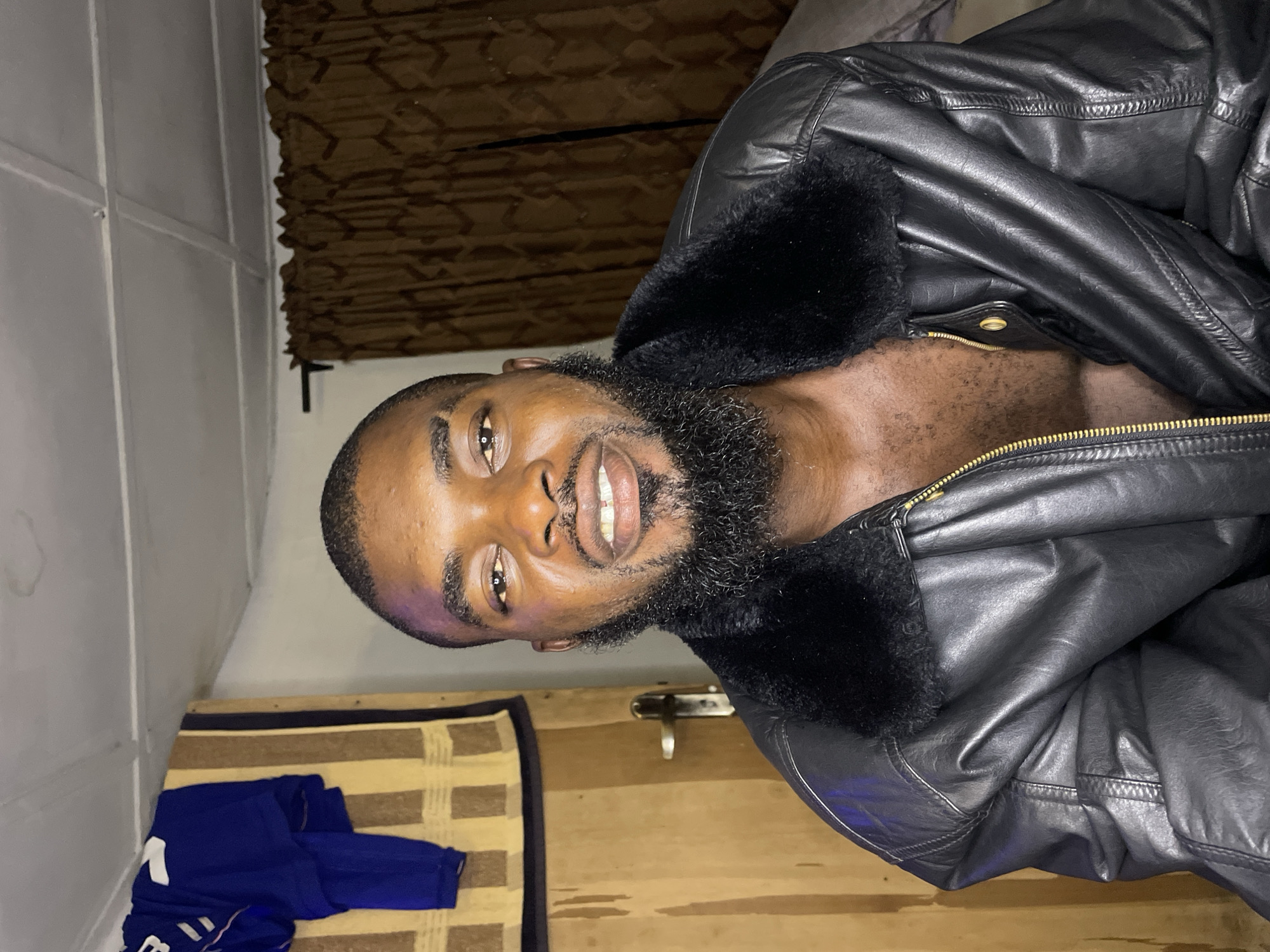 A male individual, appearing to be of Black ethnicity and approximately 20-35 years of age, is centered in a portrait shot, looking directly at the camera with a slight smile. He has short, dark hair with faded sides, a full, dark beard, and dark eyes. A faint purplish-blue light reflection is visible on his forehead above his left eyebrow. He is wearing a black leather jacket with a black faux fur collar; the jacket is unzipped, revealing his upper chest which has dark body hair. His mouth is slightly open, showing some upper teeth, including visible gaps between the central incisors.
The setting is an indoor room. To the viewer's left, a light brown wooden door with a silver-colored handle is visible. Over the door or an adjacent structure, a blue garment with white markings (possibly a sports jersey) is draped over a vertically striped brown and tan fabric. Above this, the ceiling appears white with visible structural lines or beams. To the viewer's right, dark brown curtains with a repeating geometric pattern are present. The lighting is artificial and relatively dim. No specific visual identifiers within the photograph allow for confirmation of an exact location within the city of Jos, Nigeria.