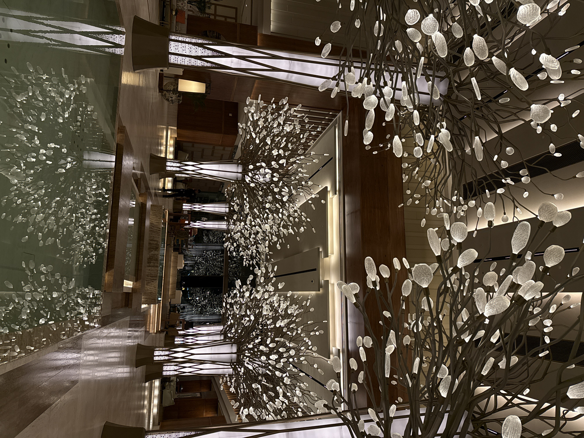 The image depicts the interior of what appears to be a hotel lobby in Dubai, United Arab Emirates. The scene is illuminated with a warm, artificial light. The architectural design is modern with repeating vertical elements. Several decorative lighting fixtures resembling stylized trees with numerous illuminated leaves or buds are prominent. These fixtures are a focal point, repeated along the length of the lobby. To the left, there is a body of water, reflecting the lights and elements of the ceiling, creating a mirror effect. The floor is polished and reflects the light and the interior elements. The space seems large and well-maintained. The absence of people suggests a quiet or off-peak time.