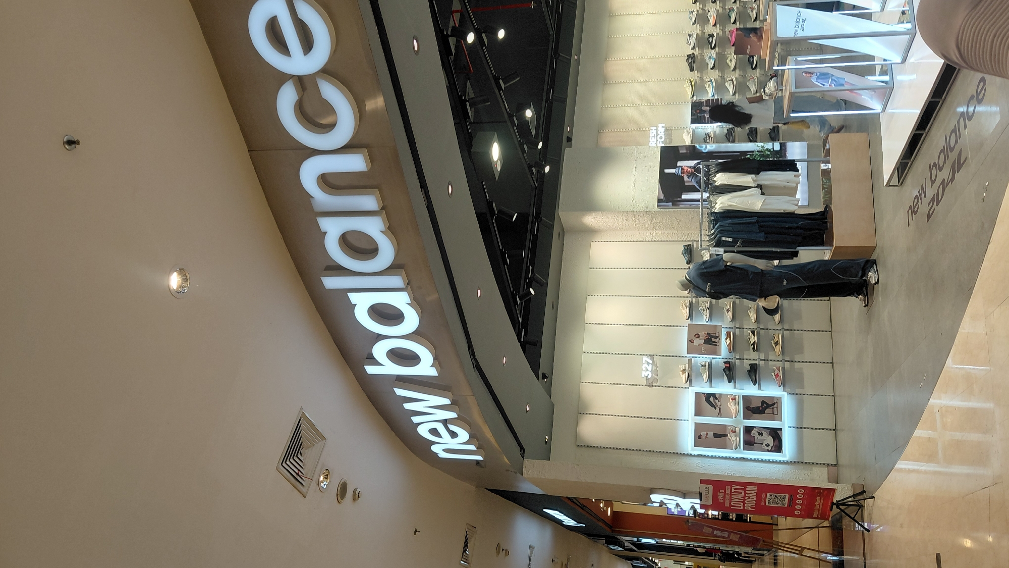 The image shows the interior of a New Balance store, likely located in the city of Purwokerto, Indonesia, as indicated by the "New Balance 2024" text on the floor and the overall mall-like aesthetic.
The store's facade features a prominent, illuminated "new balance" logo. Inside, shelves display a variety of New Balance footwear, with many pairs presented in a grid-like arrangement on the walls. A mannequin dressed in New Balance apparel stands near a rack of clothing, which includes t-shirts and jackets. In the background, a woman with long dark hair and a light-colored shirt is browsing the merchandise, and another person, partially visible, is also in the store.
A red promotional stand with the text "MA.CLUB LOYALTY PROGRAM" is positioned to the left of the entrance. The flooring is a combination of polished light-colored tiles and a grey concrete-like material. The ceiling is a light color with recessed lighting and air vents. The overall impression is a clean, modern retail environment designed to showcase athletic footwear and apparel.