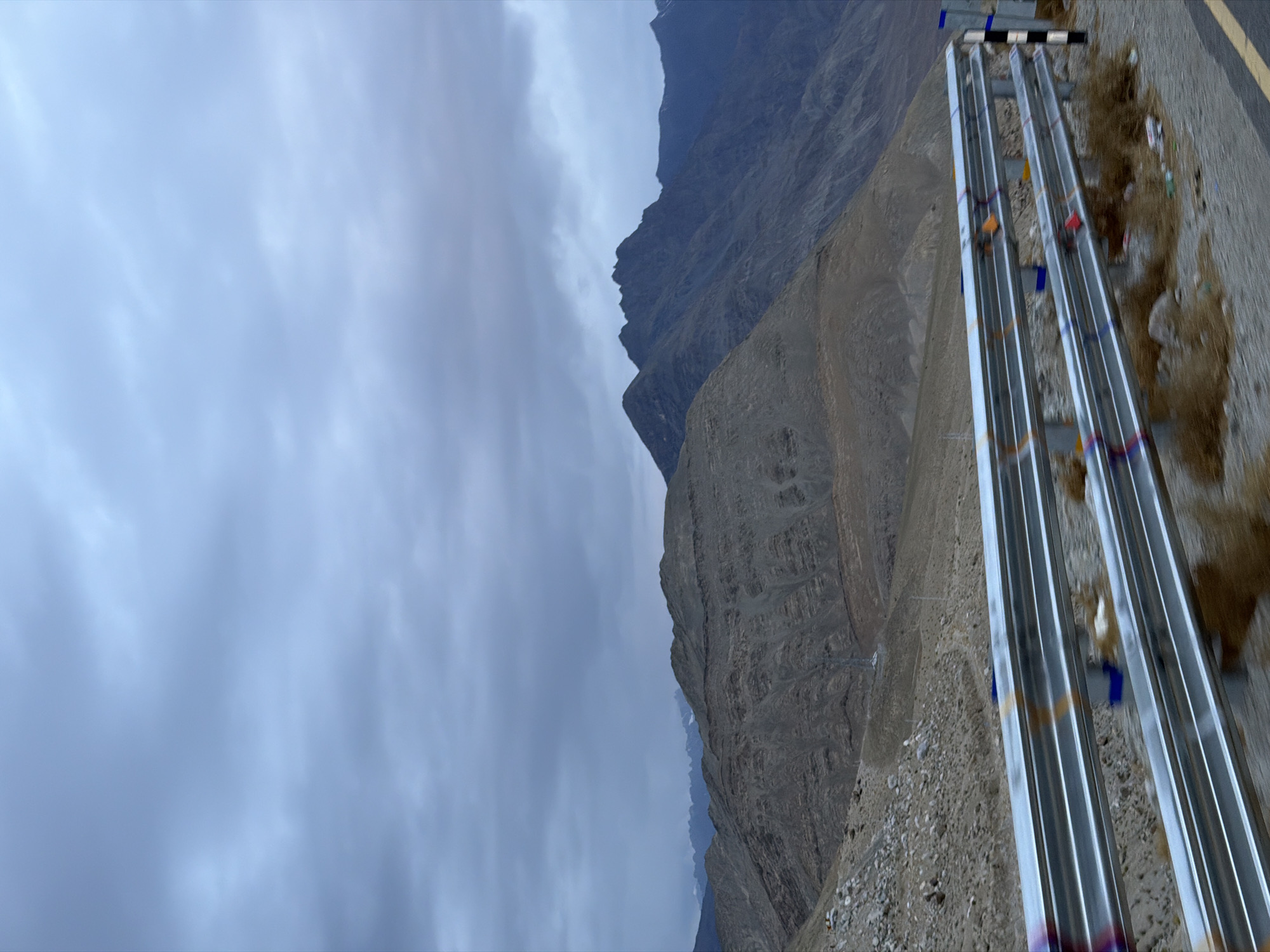 The media file depicts a mountainous landscape in Chumkar, India, under a cloudy sky. The scene is characterized by rugged, barren mountains with visible geological stratification. In the foreground, a road is visible, bordered by a series of metallic guardrails or pipes. These structures are somewhat reflective and appear to be painted with colorful ribbons or flags at intervals, suggesting a decorative or symbolic purpose, possibly related to local traditions or a significant route.
The ground next to the road is covered in gravel and sparse, dry vegetation. There are also some pieces of litter scattered along the roadside, indicating human presence. The mountains in the background are imposing and dominate the upper portion of the frame, with sharp ridges and varied textures of rock and soil. The sky is overcast, casting a diffused light over the entire scene, and hints at potentially cooler weather.
The perspective suggests the photo was taken from a slightly elevated position, looking down and across the landscape. The absence of people or a clear activity suggests a static scene, possibly a roadside view captured during transit. The overall mood is serene and somewhat stark, characteristic of high-altitude, remote environments. No visible text is present in the media file.