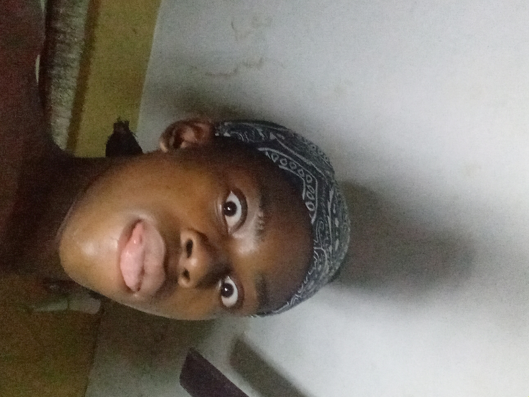 The image shows a dark-skinned person wearing a blue and white patterned bandana, with their tongue slightly protruding from their mouth. The person is looking directly at the camera. The background is a mottled white and yellow wall. The image was purportedly taken in Nkpor, Nigeria.