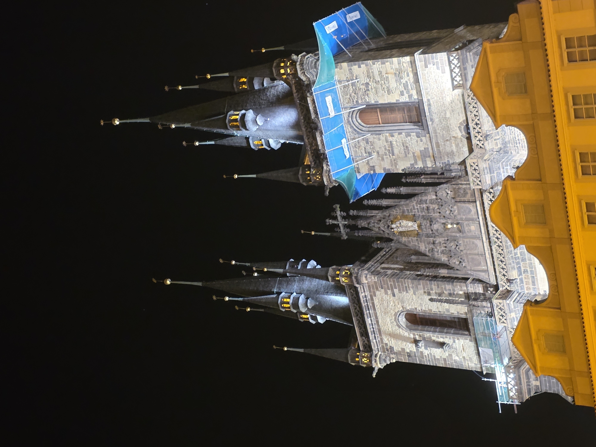 The image displays the Church of Our Lady before Týn in Old Town Square, Prague, Czech Republic, captured at night. The prominent Gothic church dominates the upper portion of the frame, illuminated against a dark, clear sky. Its two main towers rise with multiple slender spires, each culminating in a golden, star-shaped finial with a luminous spherical element beneath. Small, arched windows within the spires emit a warm, golden light. The stone facade of the towers is primarily illuminated by cool, blue-white spotlights, creating a contrast with the warmer-toned windows.
A section of the right tower is partially covered by blue and green netting affixed to scaffolding, indicating ongoing maintenance. Intricate stone carvings, including a central statue resembling the Madonna and Child, are visible in the gable between the main towers. Beneath the church, a lower building with a warm, yellow-orange facade is partially visible. This building features distinctive gabled roofs adorned with scrollwork and multiple windows fitted with closed shutters. Additional scaffolding is present on the lower left side of the church's base, positioned above the yellow building. No people are visible in the scene.