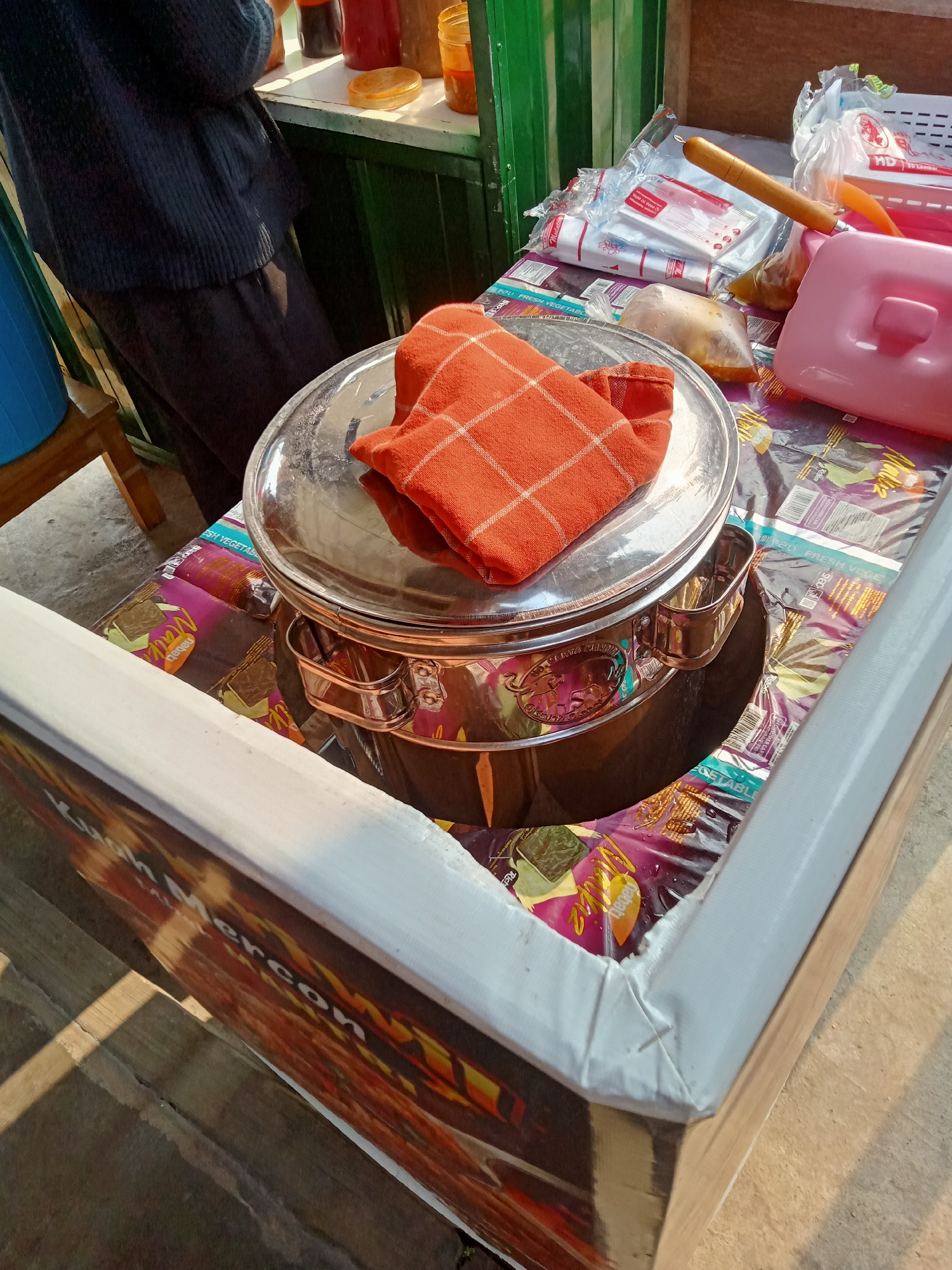 In Simo, Indonesia, a street vendor operates a small food cart. The cart is constructed of wood and is lined with a white material on the top edge. The cart's surface is covered with colorful, patterned plastic sheeting. On the cart, a large, shiny metal pot with a lid is prominent. An orange checkered cloth is folded on top of the lid. To the right of the pot, there are several items including a pink plastic container with a handle, a bag of food, and a wooden utensil with a metal end. In the background, a person wearing a dark blue knitted sweater and dark pants stands behind a counter. On the counter, there are bottles of liquid and a yellow object. To the left of the cart, a blue plastic container is visible. The scene is lit by sunlight, casting shadows on the ground.