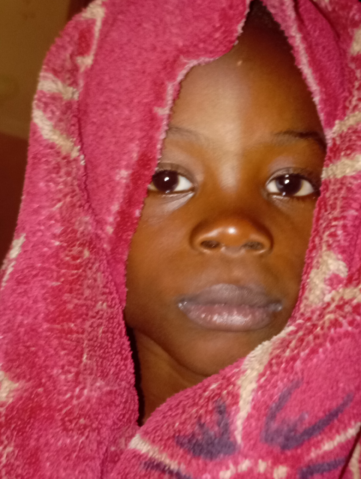 The image shows a close-up of a young child's face, wrapped in a red and cream patterned towel or blanket. The child's eyes are open and looking towards the camera. The towel covers the child's head and wraps around their face, obscuring most of their features except for their eyes, nose, and mouth. The background is out of focus and appears to be a dimly lit interior space.