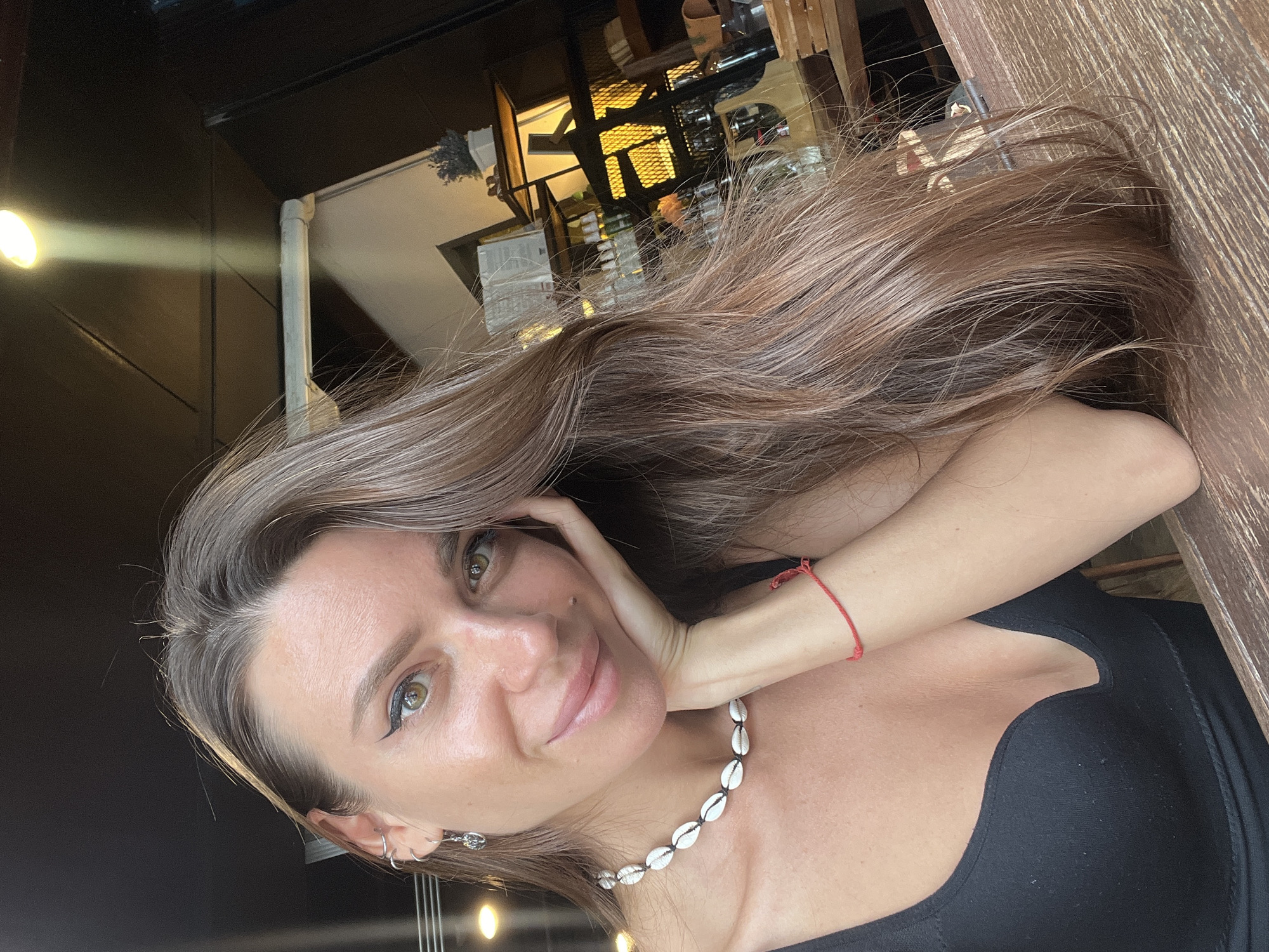 A young Caucasian woman is pictured from the chest up, resting her head on her right hand, which is propped on a wooden surface. She has long, light brown hair that falls over her right shoulder and partially obscures the background. She is wearing a black, low-cut top and a necklace made of white shells. A thin red string bracelet is visible on her left wrist. Her left ear has two silver hoop earrings and a stud. The background appears to be an indoor setting, possibly a cafe or restaurant, with shelves of bottles and a warm light source illuminating the scene. The lighting is soft and natural, casting subtle shadows. The woman is looking directly at the camera with a slight smile. The overall tone of the image is casual and relaxed.