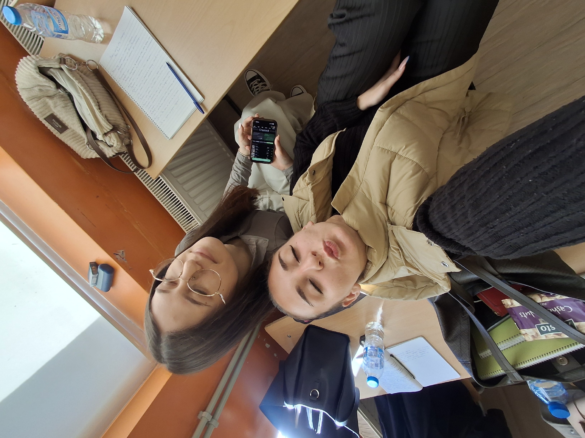 Two young women are positioned at adjacent desks in what appears to be an academic environment, possibly a university or school classroom. One woman wears glasses and has dark hair, while the other is wearing a tan puffer jacket. Both have their eyes closed. A phone displaying what appears to be a user interface of a social media platform is being held by the woman with dark hair. Items such as notebooks, pens, water bottles, and bags are scattered on the desks. The setting could be in Sofia, Bulgaria given the possible academic setting and general aesthetic.