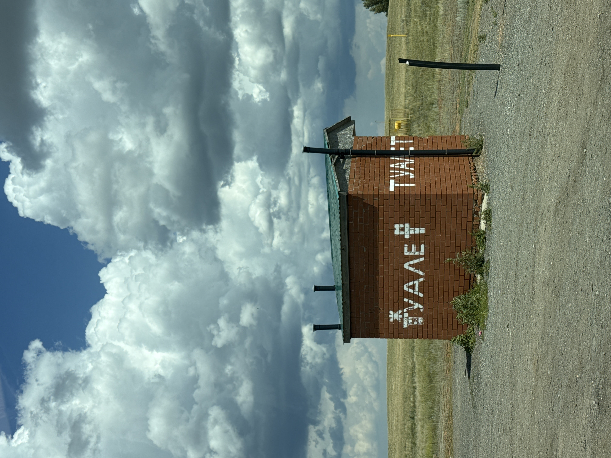 The image shows a brick outhouse with a green roof situated in an open, grassy field under a dramatic sky filled with large, grey clouds. The outhouse has white lettering on its brick facade that reads "ТУАЛЕТ" on one side, which translates to "TOILET" in English. Two dark pipes, likely chimneys or vents, protrude from the roof. The ground in front of the outhouse is covered with gravel. In the background, the field extends to the horizon, with some yellow markers and a fence visible in the distance. There are no people or animals present in the image. This scene is consistent with an outdoor facility in a rural or undeveloped area, possibly on the outskirts of Pervomaiskii, Russia.
