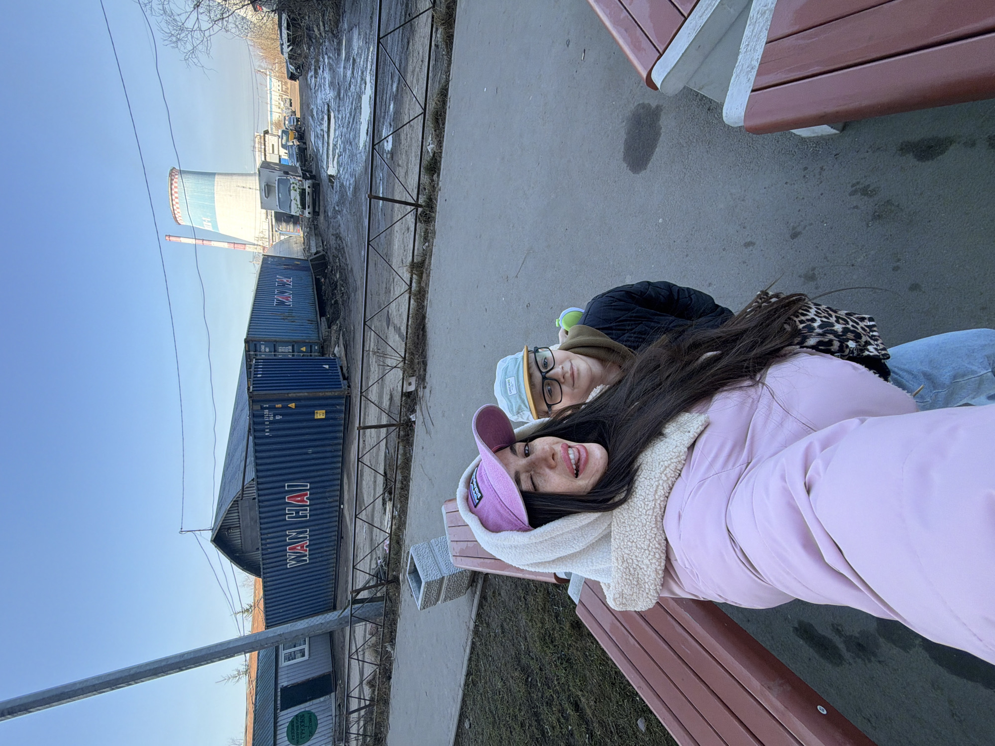 The image captures a wide-angle selfie featuring two individuals in an industrial area of Saint Petersburg, Russia, specifically near Thermal Power Plant 19 (ТЭЦ-19) at Prospekt Narodnogo Opolcheniya, 137. The prominent cooling tower of ТЭЦ-19, marked with "ТЭЦ-19" text and distinguished by white, light blue, and red bands at its upper section, is visible in the background.
In the foreground, a female individual with long dark hair is seated on a brown wooden slatted bench. She wears a pink puffer jacket over a leopard-print garment, light blue jeans, and a pink baseball cap displaying the Patagonia logo. Her right eye is winked, and her tongue is extended slightly, directed towards the camera. To her right, partially visible, a second individual, a male child, is present. He wears a dark quilted jacket, a brown hoodie underneath, a light blue baseball cap with "CHICAGO" text, and black-framed glasses, looking towards the camera with a neutral expression. A green object is held in his right hand.
The immediate environment includes a grey asphalt pathway with brown wooden benches and a square concrete trash receptacle. Beyond the pathway, a low, dark metal fence with a diamond pattern separates the foreground from an unpaved, muddy industrial yard. This yard contains several large blue shipping containers, one of which prominently displays "WAN HAI" in white lettering. Behind these containers, a small light-grey building with a green sign and an older wooden structure with a curved roof are visible. A line of vehicles, including a white box truck, is parked on the muddy ground. The scene is set under a clear, bright blue sky, with sparse, bare tree branches visible on the far right.