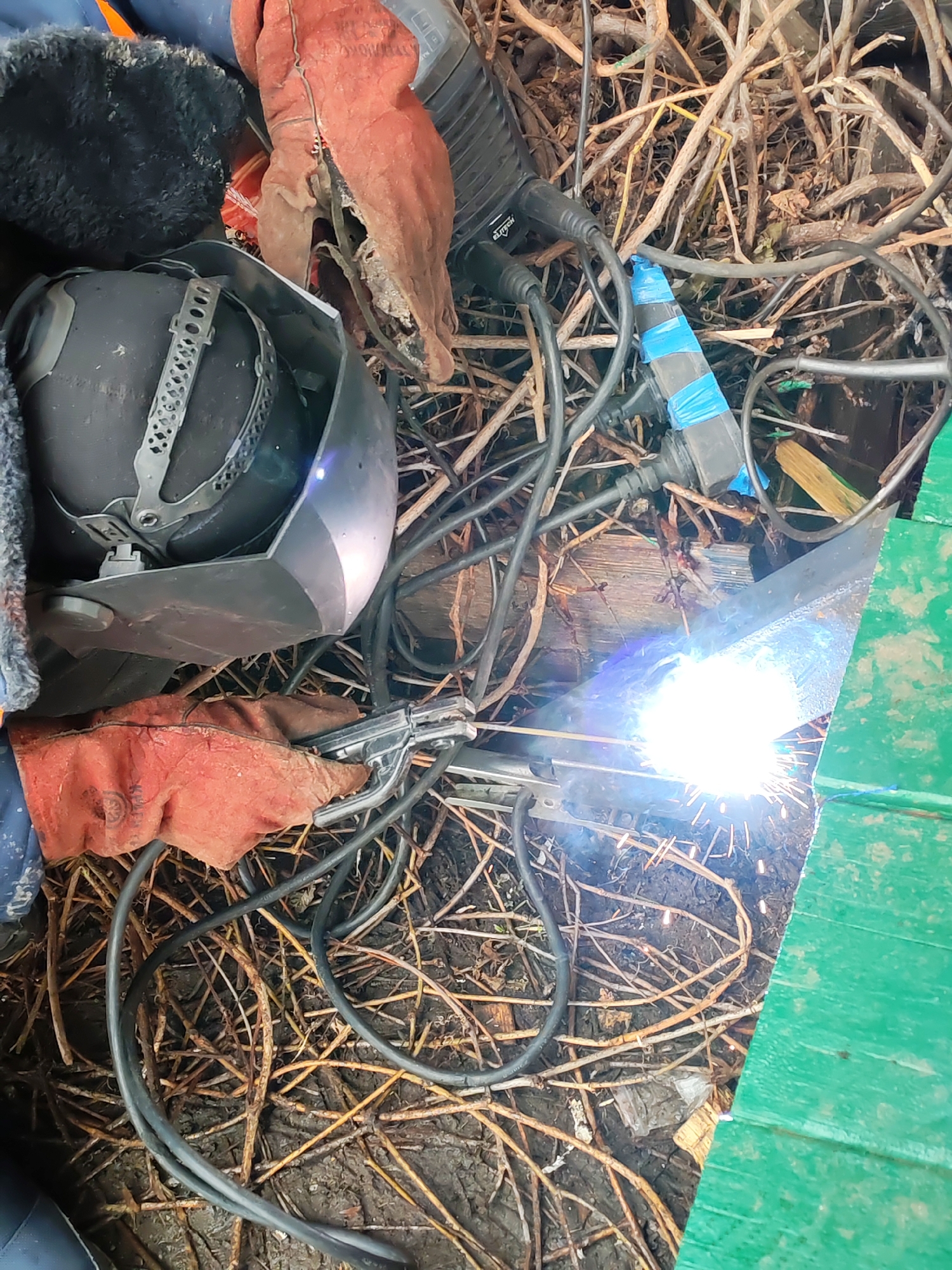 The image shows a welding operation in an outdoor environment, possibly in Chaikovsky, Russia. A welding helmet and a pair of red welding gloves are positioned near the welding area. Welding equipment, including cables, is visible. A bright arc indicates active welding. The background consists of dirt, dry twigs, and what appears to be a green painted surface.