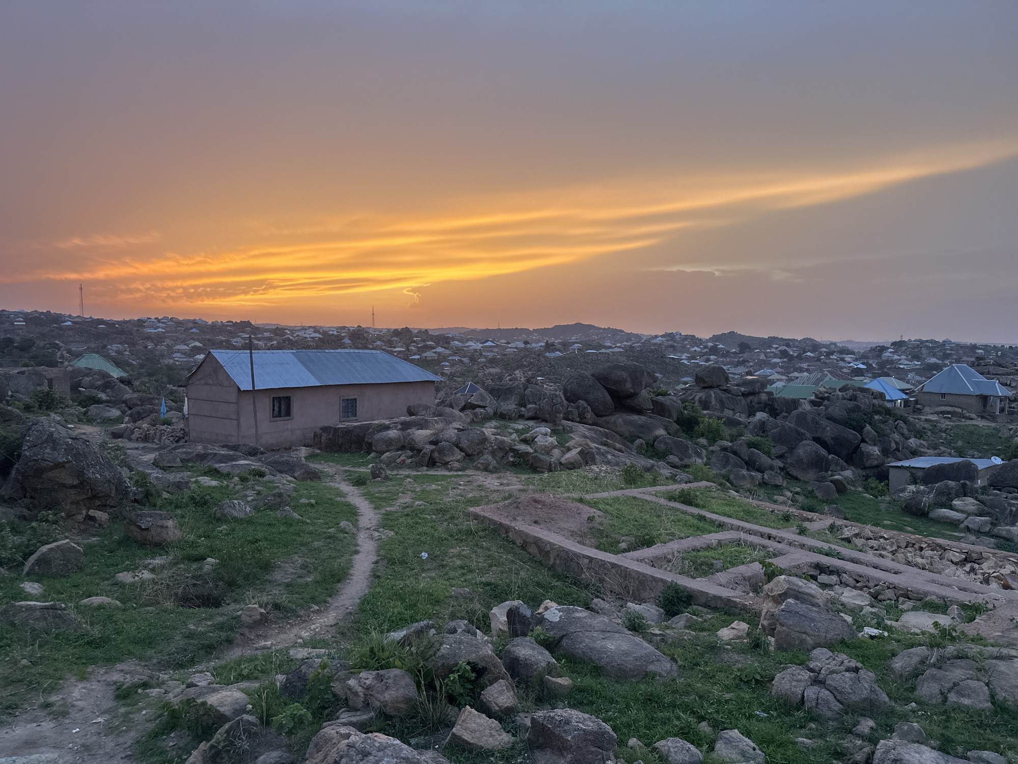 The image depicts a landscape at dusk in Jos, Nigeria. The foreground shows a rocky terrain with scattered boulders and patches of green vegetation. A narrow dirt path winds toward a small, rectangular building with a corrugated metal roof and a single window visible. Nearby are partial foundation walls. The middle ground features numerous similar buildings interspersed with rock formations, forming a dense settlement that extends to the horizon. The background is dominated by a dramatic sky gradient, transitioning from a vibrant orange near the horizon to a muted gray-blue higher up. Some tower masts are visible on the horizon.