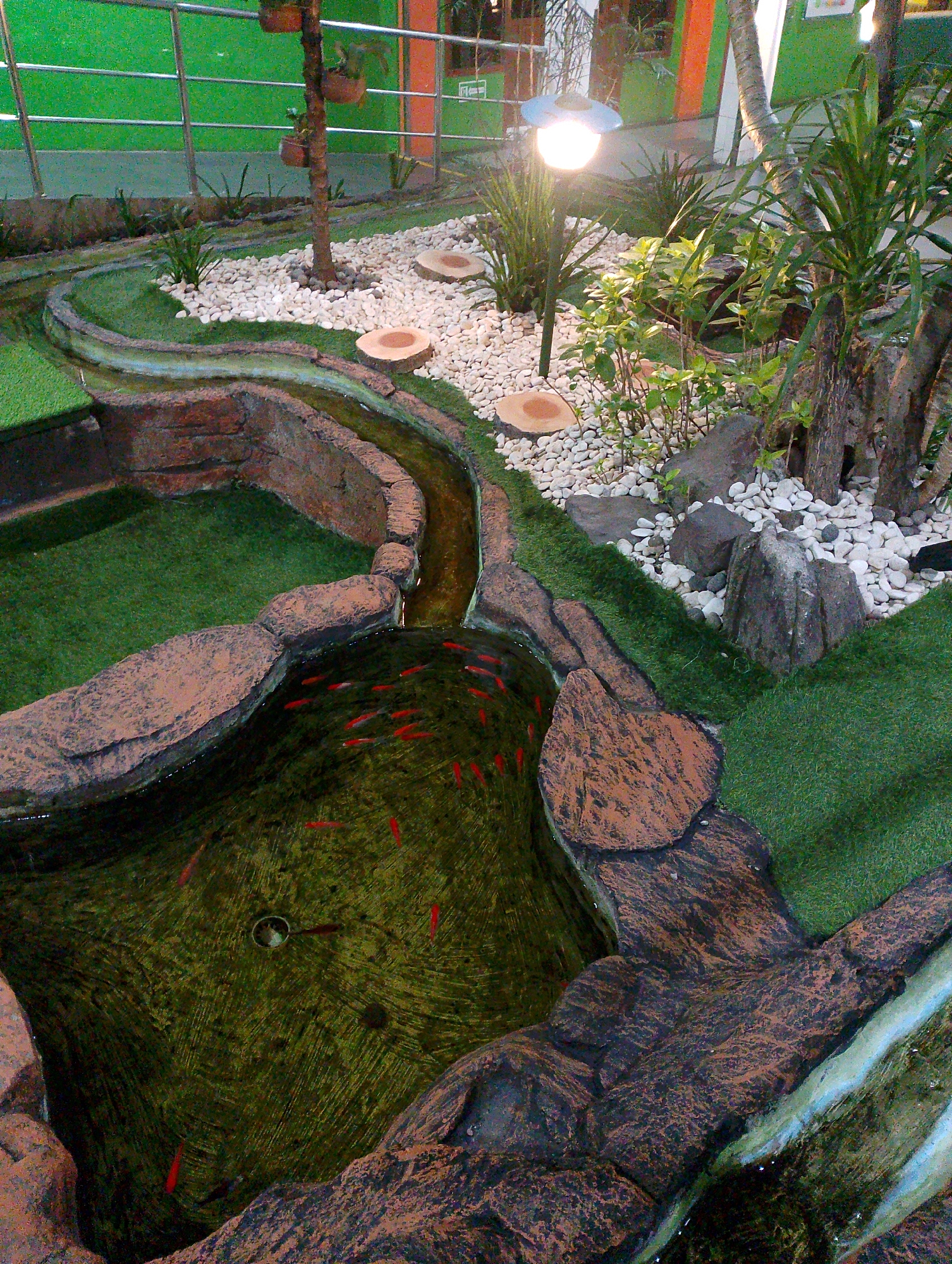 The image depicts a man-made garden with a water feature in Sidoarjo, Indonesia. A winding stream, bordered by artificial rock formations and artificial grass, leads to a pond. The pond is populated with several small red fish. The surrounding area is adorned with white pebbles, various plants, and a small tree with hanging planters. A lamppost with a bright light is also visible. In the background, a green wall and railings suggest the garden is part of a larger facility, possibly a school or public space. The overall impression is one of a decorative and tranquil garden setup.