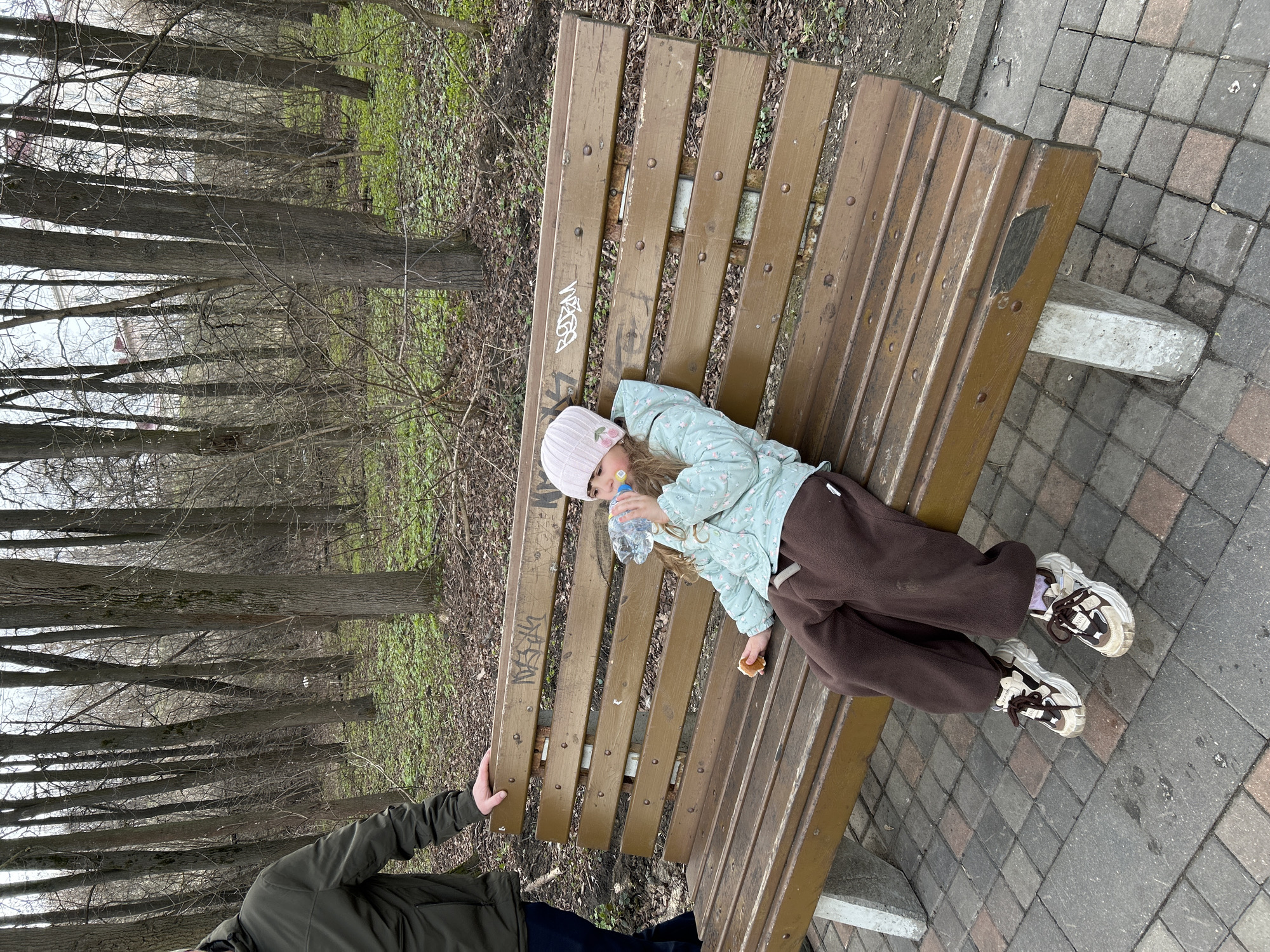 A young girl lies on a wooden bench in what appears to be a park or green area in Klin, Russia. The bench is situated on a paved area of gray and reddish bricks. In the background, a grove of bare trees can be seen. The girl is wearing a pink knit hat with flower appliques, a light blue quilted jacket, and brown sweatpants. She is holding a plastic bottle of water in her right hand and what appears to be a piece of bread in her left. A person with dark clothing is partially visible on the left side of the photo, touching the bench.