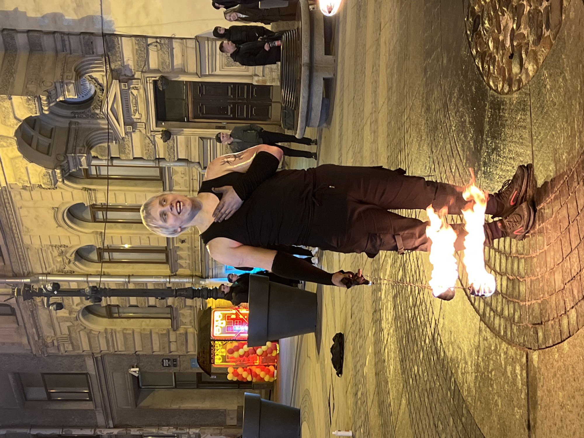 The image depicts a street performer in Saint Petersburg, Russia, engaging in a fire performance. The performer, with short white hair and wearing all black clothing, is holding a flaming object near a circular metal structure on the ground. In the background, there is a building with detailed architectural features and decorative elements. A small crowd is observing the performance. Other details include planters, an illuminated sign and strung balloons, and other people walking on the sidewalk.