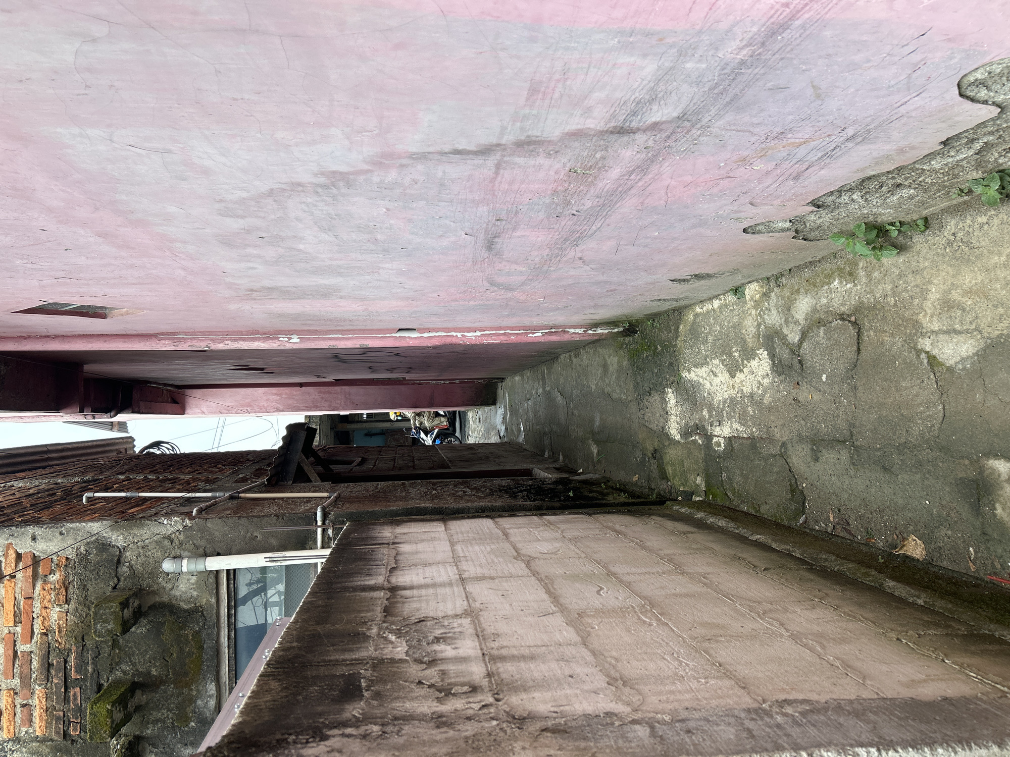 The image shows a narrow alleyway between two buildings. The perspective is looking upwards from the ground level. One building has a pink-colored wall, showing signs of wear and tear with cracks and scuff marks. There's a small, square opening in this wall. The other building is constructed with brick and has a tiled roof. A white pipe runs horizontally along the exterior of the brick building.
Further down the alley, there's a glimpse of a motorcycle parked, suggesting that this is a functional alleyway. The ground surface in the foreground appears to be made of concrete slabs. The overall impression is one of a densely built urban environment, possibly in a residential area. The lighting suggests it could be daytime, with the sun casting shadows, though no specific time can be determined. There are no visible people in the immediate foreground, but the presence of the motorcycle implies human activity. The weather appears to be fair, with no indication of rain. The location context is East Jakarta, Indonesia, which aligns with the architectural style and urban setting. No discernible text is visible in the image.